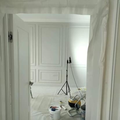 Pokój w trakcie malowania z dekoracyjnymi panelami ściennymi, widoczny sprzęt malarski i oświetlenie na statywie, podłoga zabezpieczona folią.