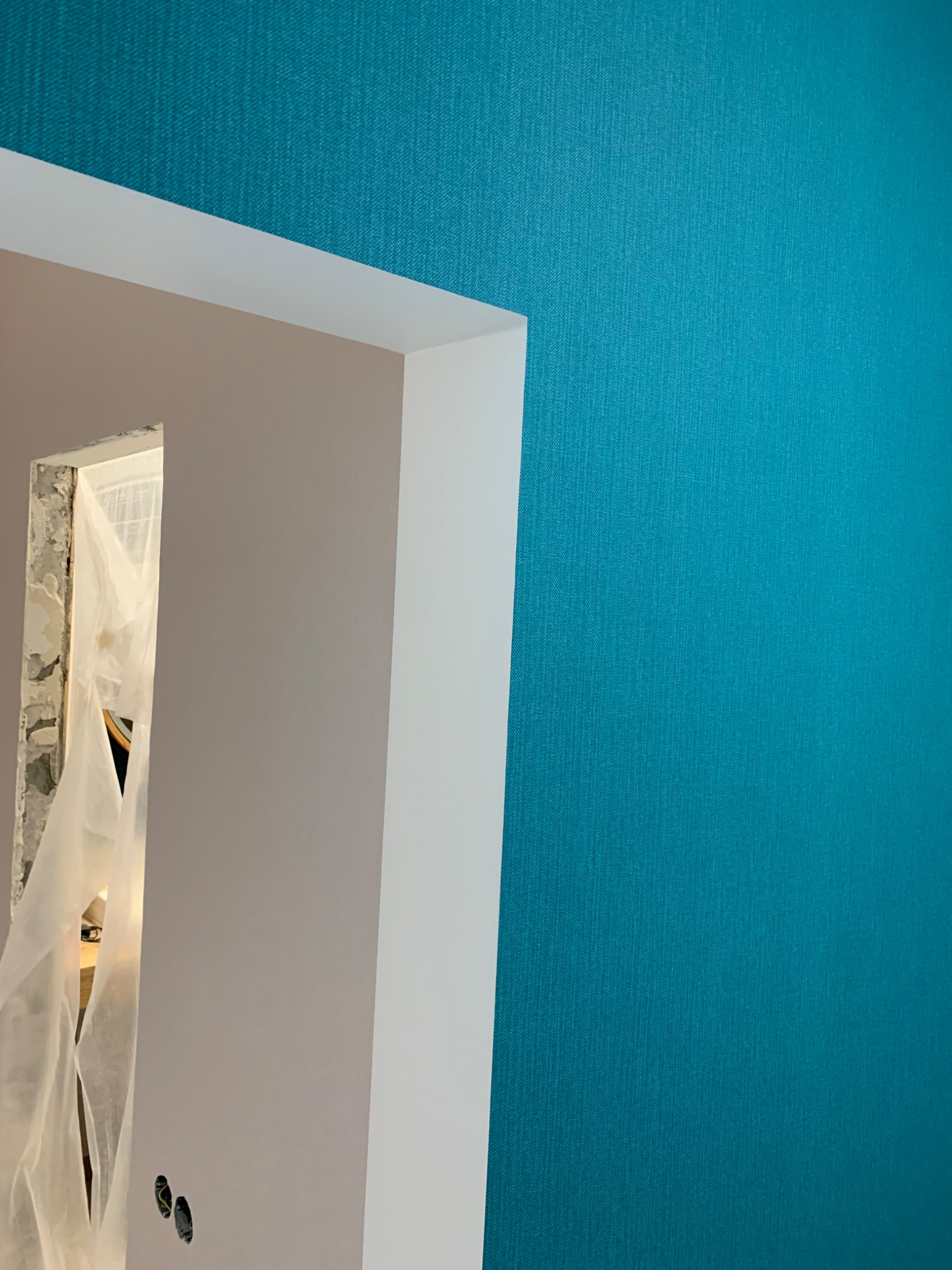 Fragment pokoju w trakcie remontu: turkusowa ściana z widoczną strukturą, pomalowana ściana w odcieniach szarości z przygotowanym otworem na gniazdka elektryczne, zabezpieczone folią drzwi w tle.