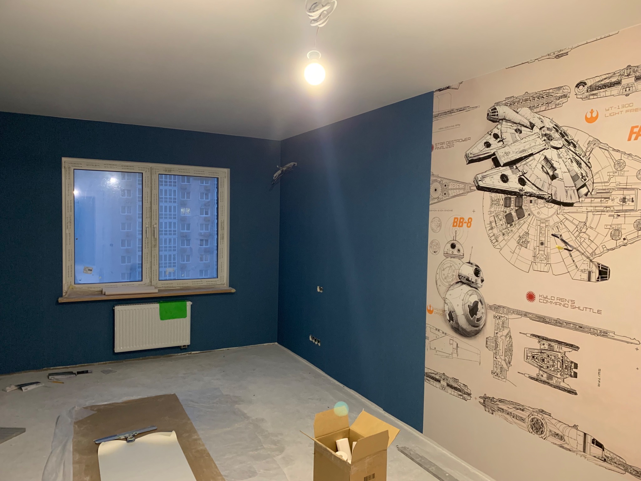Pokój po remoncie z ciemnoniebieskimi ścianami i tapetą z motywami Star Wars na jednej ze ścian. Na podłodze widoczne narzędzia i karton, a na ścianie grzejnik z zielonym zabezpieczeniem.