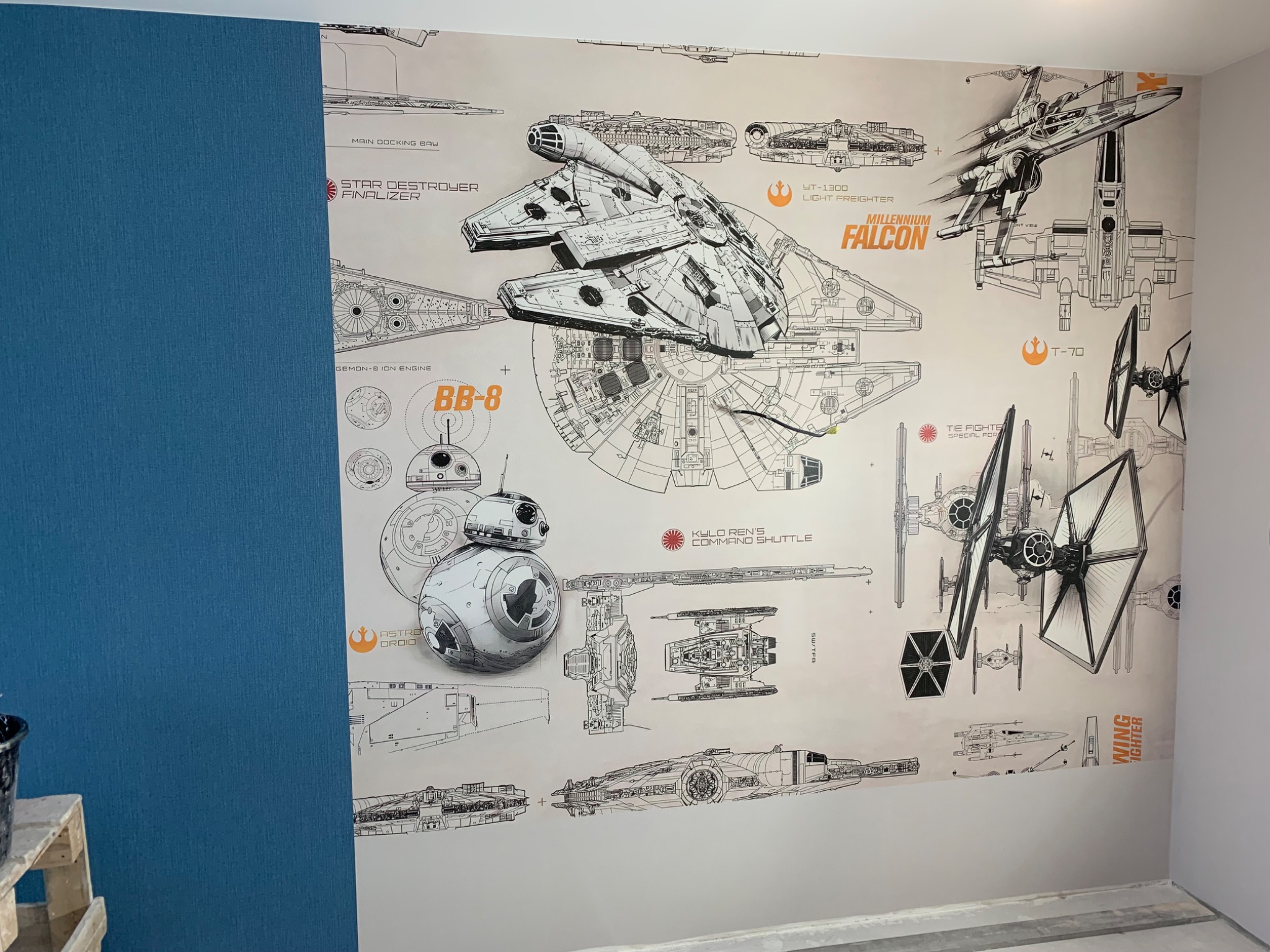 Ściana z tapetą przedstawiającą schematy statków kosmicznych z Gwiezdnych Wojen, w tym Millennium Falcon, BB-8, X-Wing i inne, częściowo zasłonięta przez pomalowaną na niebiesko ścianę z widocznym...