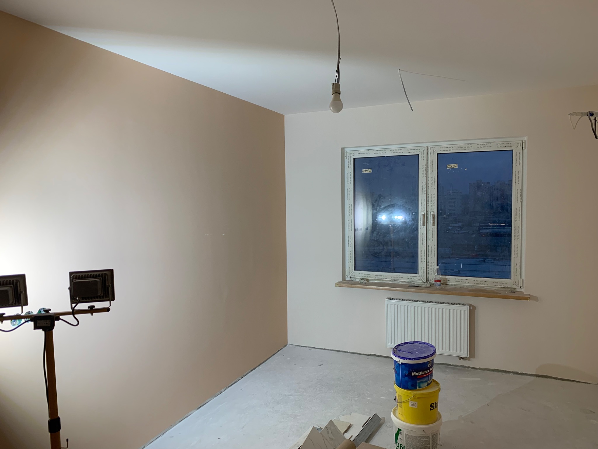 Pokój po częściowym odświeżeniu ścian w odcieniach beżu i bieli, widoczne puszki z farbą, grzejnik, okno z widokiem na miasto nocą oraz lampa budowlana na statywie