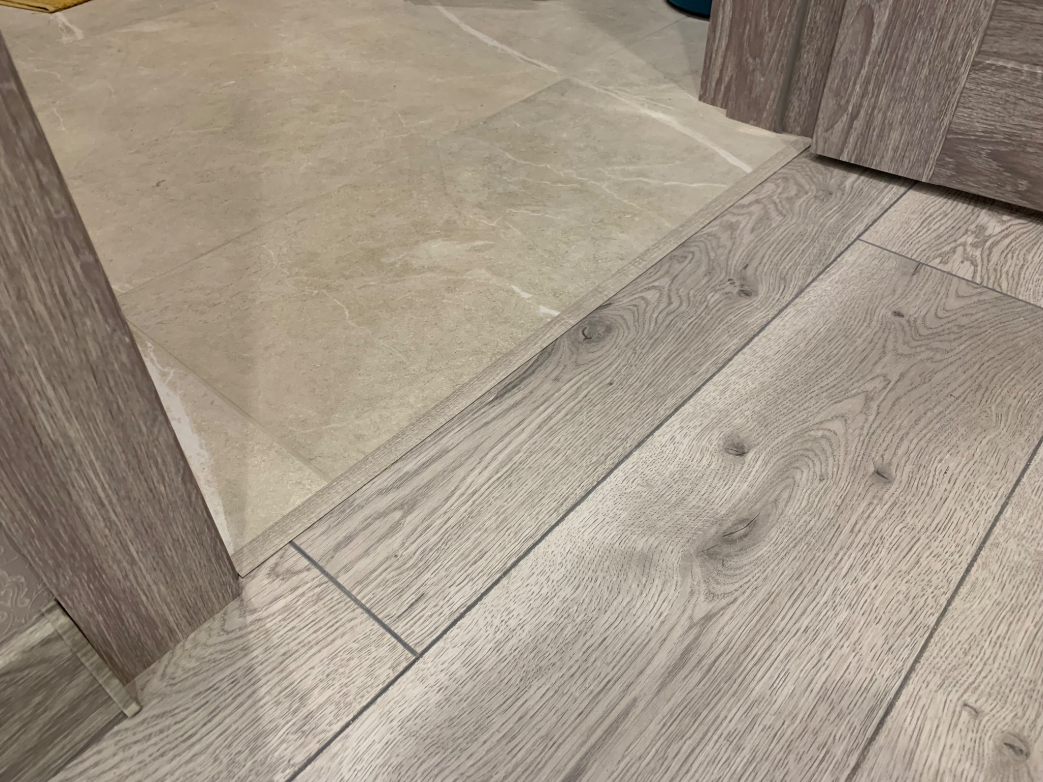 Połączenie ułożonych paneli podłogowych w odcieniach szarości z jasnymi płytkami ceramicznymi, widoczna krawędź szafki w kolorze paneli.