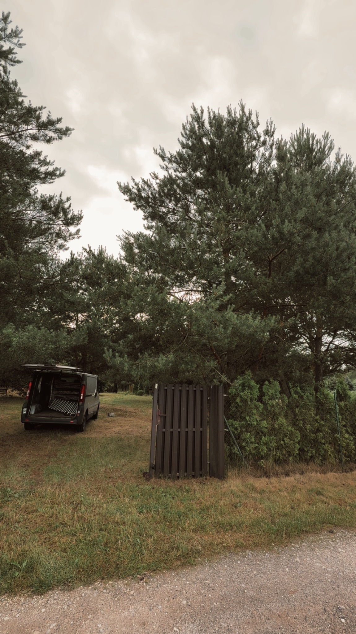 Otwarty van z elementami ogrodzenia w tle, otoczony zielenią i drzewami, sugerujący transport materiałów budowlanych lub ogrodniczych na posesję.