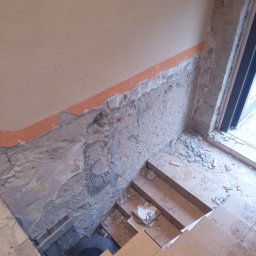 Adam Dombke gamma - Demontaż starej okładziny ściennej w klatce schodowej, widoczne fragmenty starej farby, gruz i pracownik w ciemnym ubraniu w dolnej części kadru.