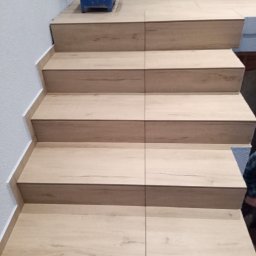 Adam Dombke gamma - Schody wykończone panelami imitującymi drewno, z widocznym niebieskim wiadrem malarskim na górnym podeście i listwą wykończeniową przy ścianie.