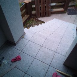 Adam Dombke gamma - Układanie szarych płytek na schodach wejściowych do budynku, widoczne narzędzia i fragment ogrodzenia.