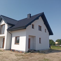 Adam Dombke gamma - Nowoczesny, dwukondygnacyjny dom z ciemną dachówką i jasną elewacją w trakcie wykańczania, widoczny fragment rusztowania i żółty samochód dostawczy w tle.