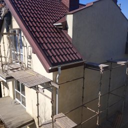 Adam Dombke gamma - Praca na wysokości: mężczyzna na rusztowaniu przy elewacji domu z brązową dachówką, widoczny fragment okna i rynny, jasna elewacja w trakcie prac.