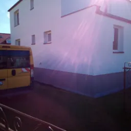 Jasny budynek jednorodzinny z nową, szarą izolacją fundamentów, widoczny zza ogrodzenia i żółtego samochodu dostawczego Fiat Ducato z numerem rejestracyjnym GPU 3MA2.