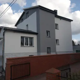 Dwukondygnacyjny dom z ocieploną elewacją w odcieniach szarości i bieli, widoczny zza ceglanego muru z czarnym ogrodzeniem.