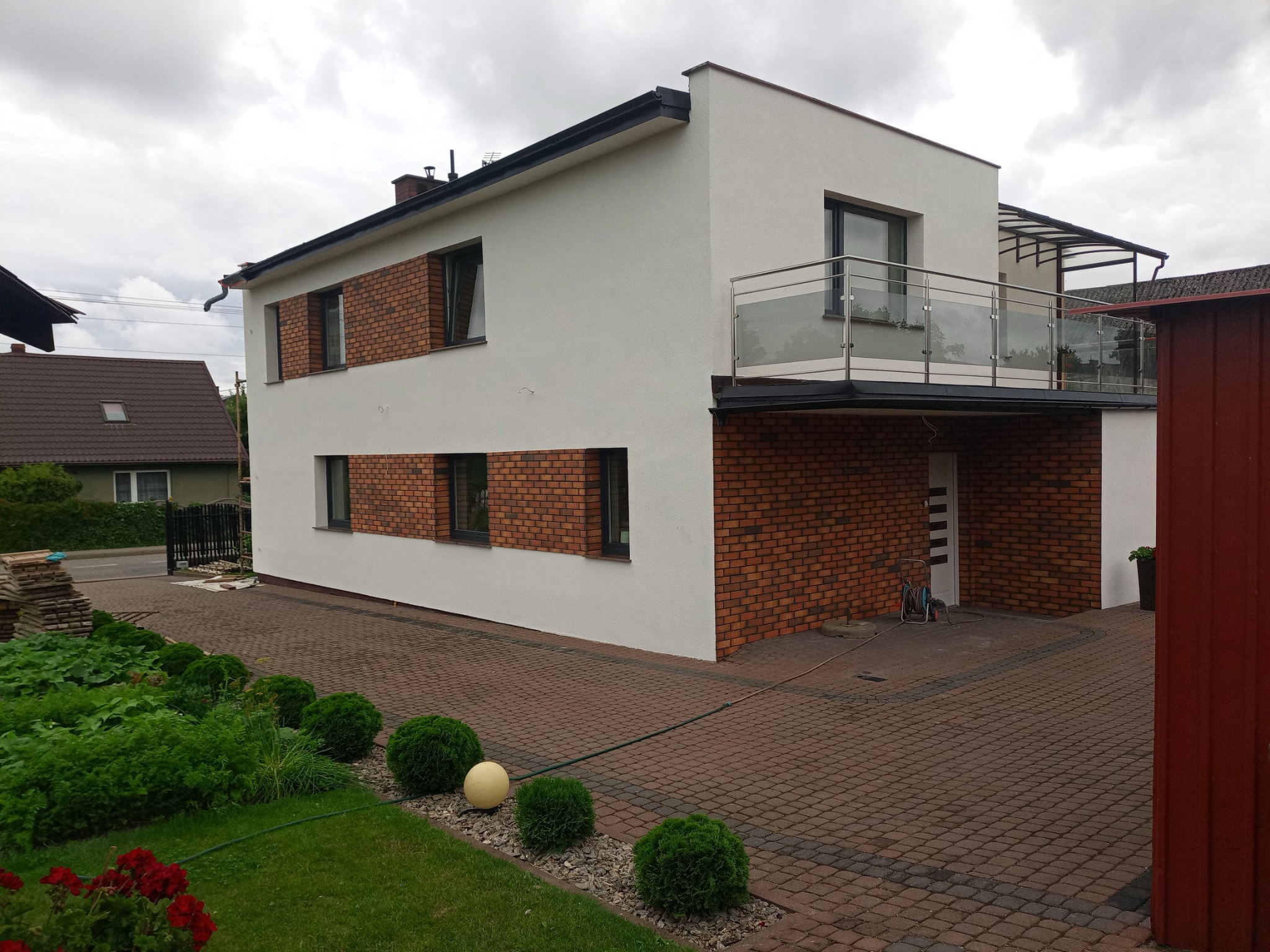Dwukondygnacyjny dom z białą elewacją i fragmentami wykończonymi cegłą, z balkonem z szklaną balustradą, otoczony zadbanym ogrodem z kulistymi krzewami i czerwoną kostką brukową.