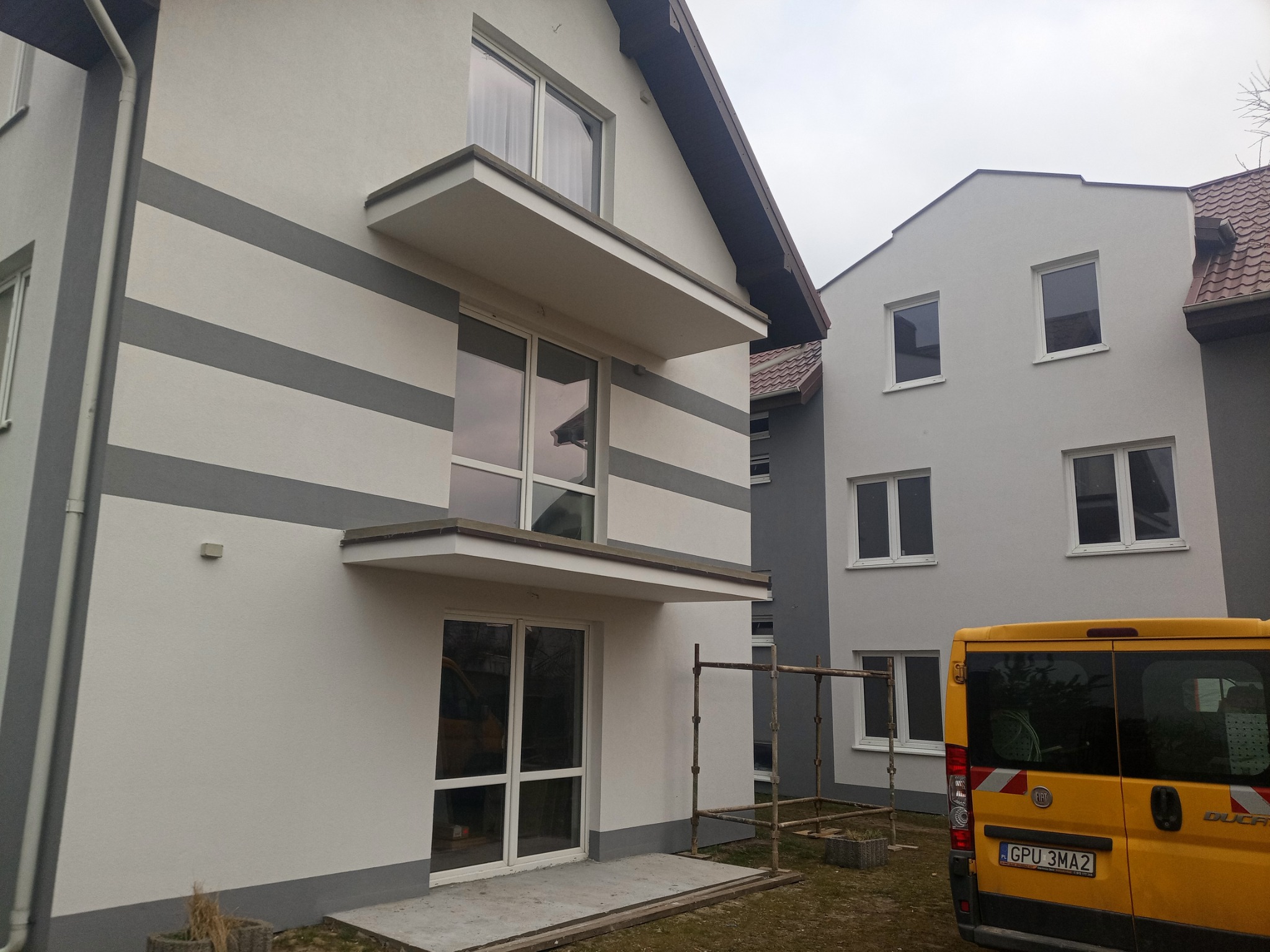 Otynkowana elewacja domu jednorodzinnego z szarymi pasami i balkonami, widoczny fragment żółtego samochodu dostawczego i rusztowanie.