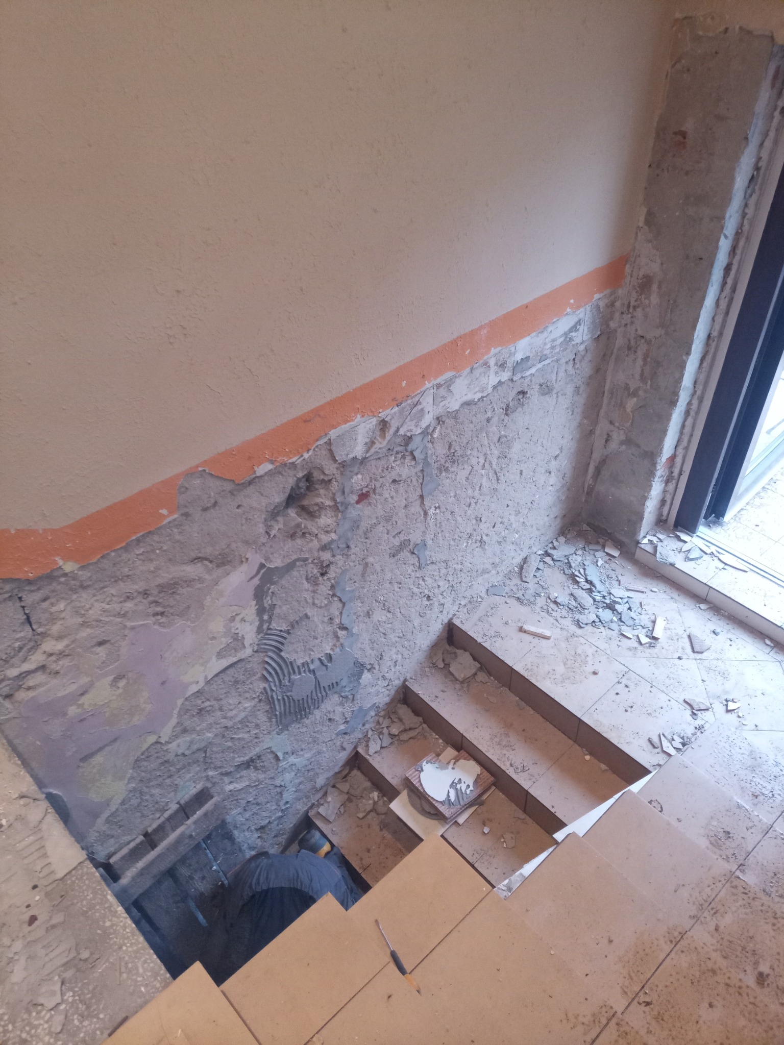 Demontaż starej okładziny ściennej w klatce schodowej, widoczne fragmenty starej farby, gruz i pracownik w ciemnym ubraniu w dolnej części kadru.