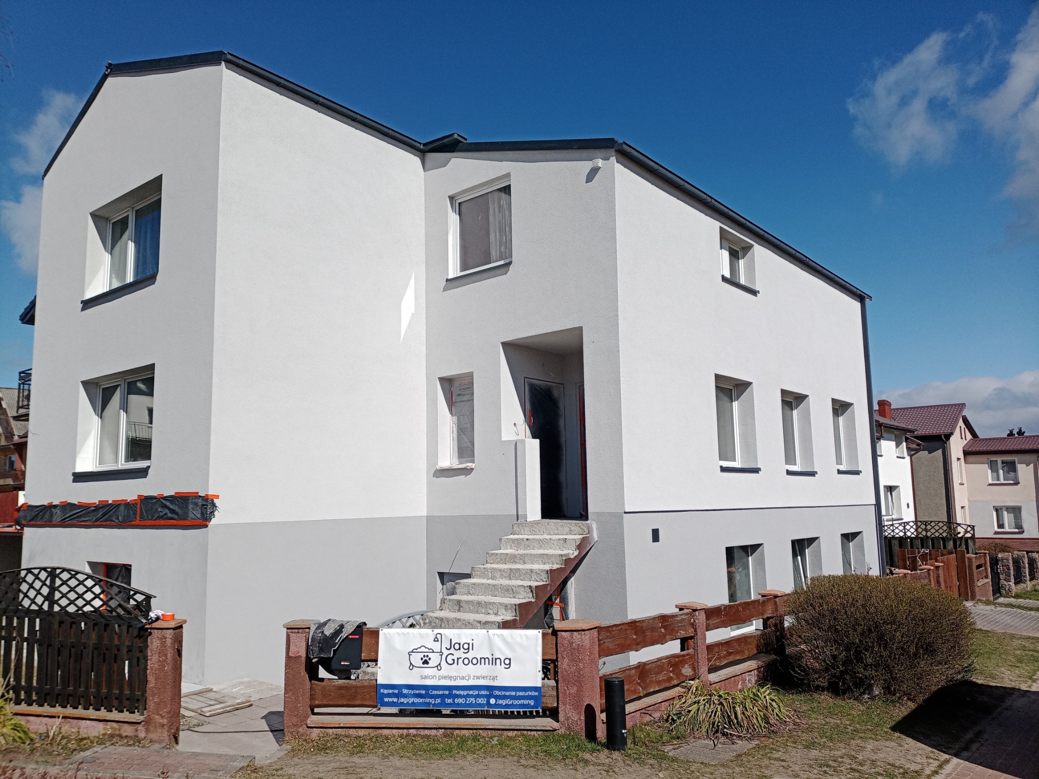 Dwukondygnacyjny budynek po świeżym malowaniu elewacji w odcieniach szarości i bieli, z widocznym zabezpieczeniem folią okien na parterze, oraz banerem salonu pielęgnacji zwierząt przed wejściem.