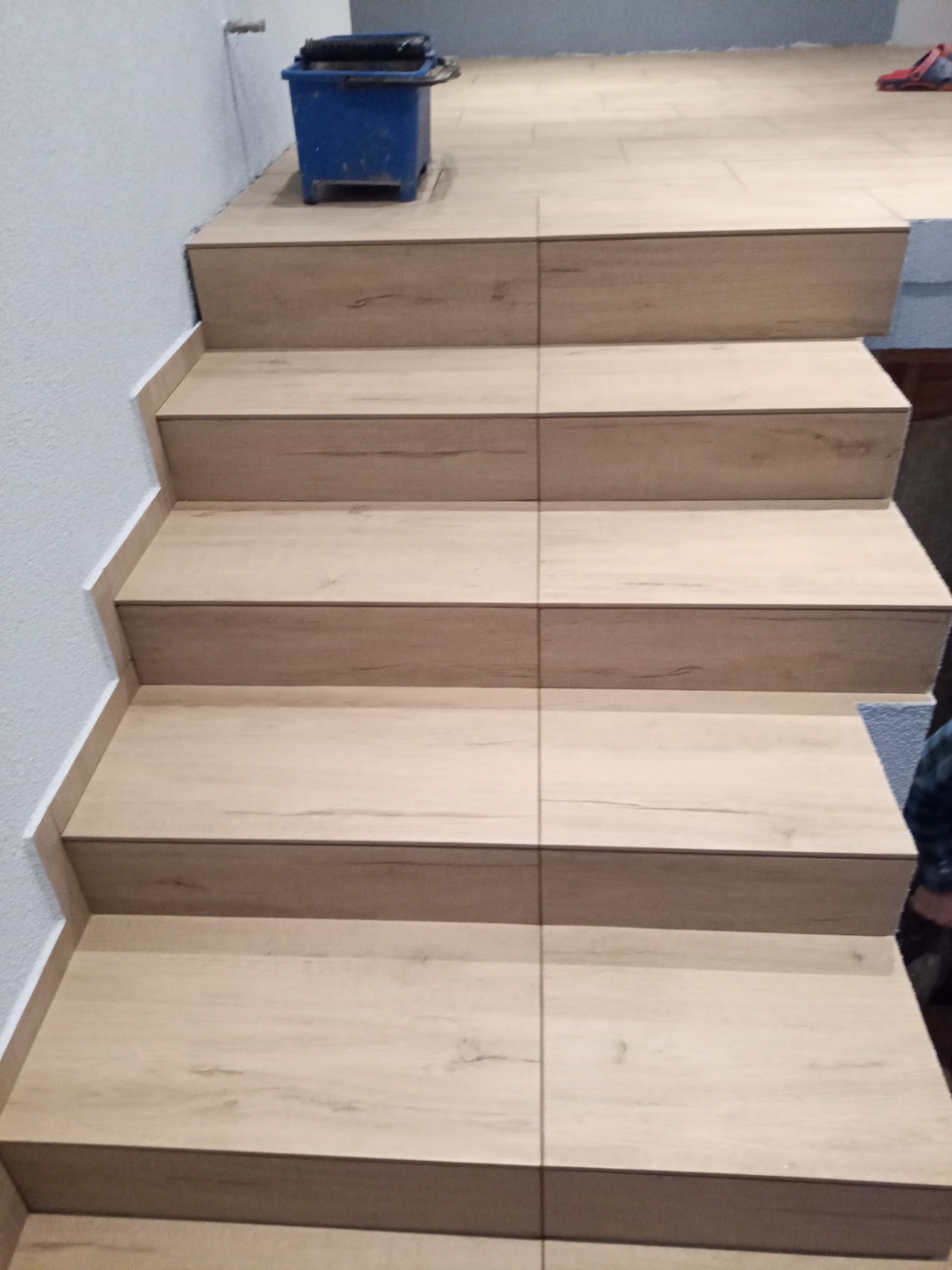 Schody wykończone panelami imitującymi drewno, z widocznym niebieskim wiadrem malarskim na górnym podeście i listwą wykończeniową przy ścianie.