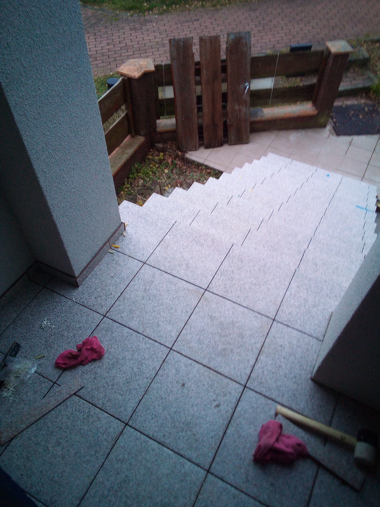 Układanie szarych płytek na schodach wejściowych do budynku, widoczne narzędzia i fragment ogrodzenia.