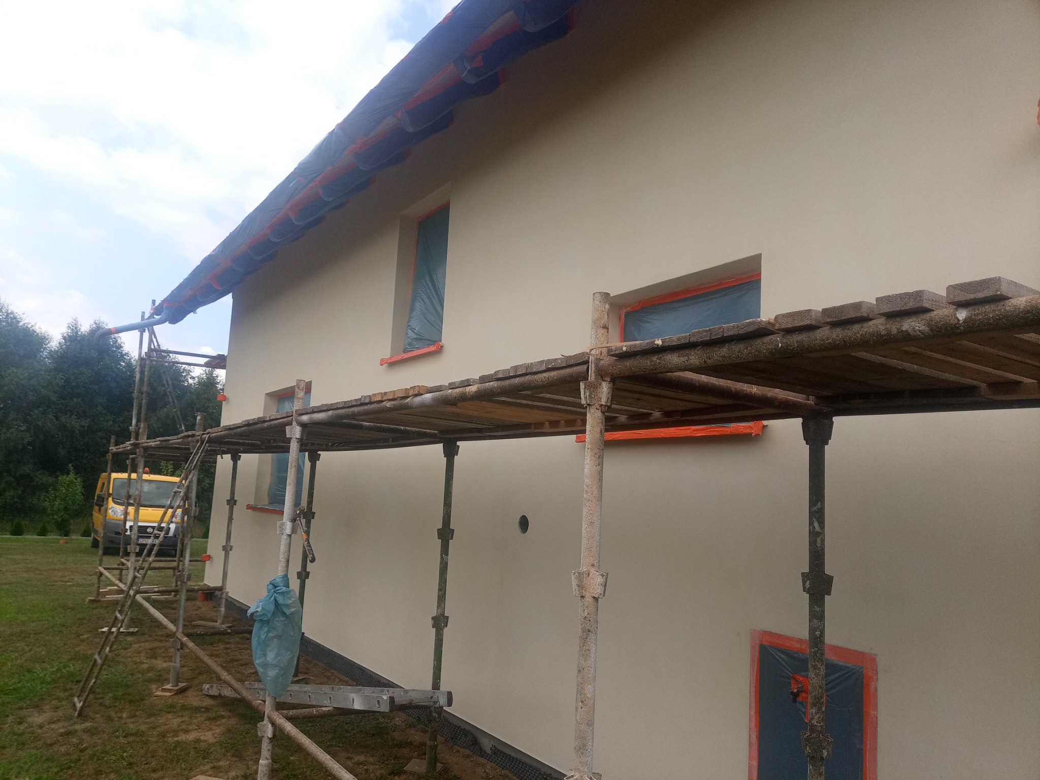 Dom jednorodzinny w trakcie ocieplania elewacji, okna zabezpieczone folią, rusztowanie z drewnianymi pomostami, żółty bus firmy w tle.