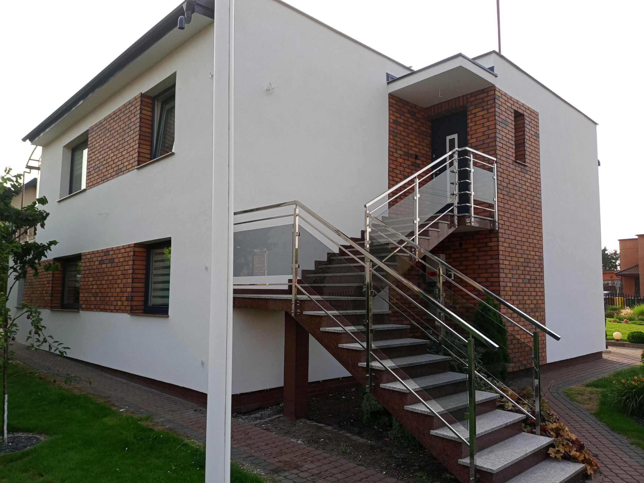 Nowoczesny dom z białymi ścianami i ceglanymi elementami, schody zewnętrzne z granitu z metalową balustradą, widok z poziomu gruntu
