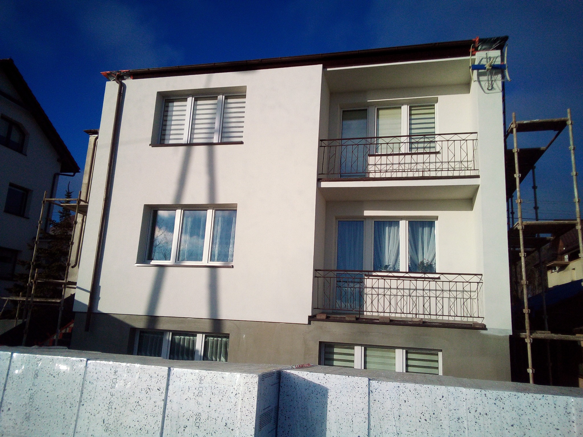 Dwupiętrowy budynek po remoncie elewacji, z widocznymi rusztowaniami i styropianem przygotowanym do dalszych prac, błękitne niebo w tle.