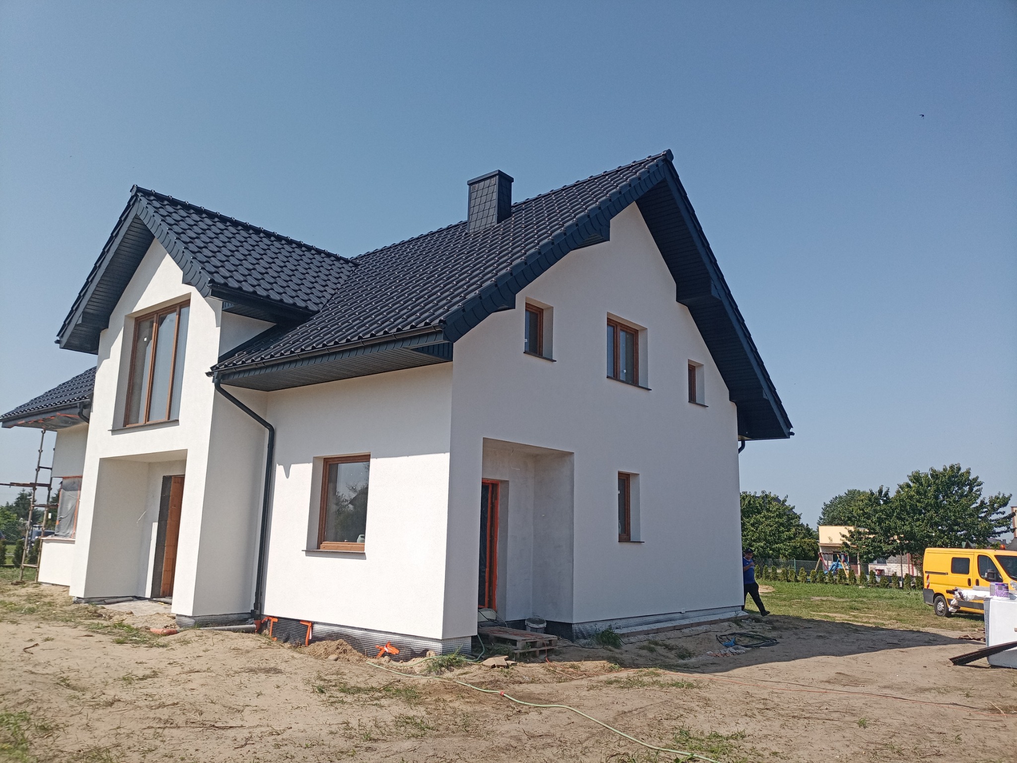 Nowoczesny, dwukondygnacyjny dom z ciemną dachówką i jasną elewacją w trakcie wykańczania, widoczny fragment rusztowania i żółty samochód dostawczy w tle.