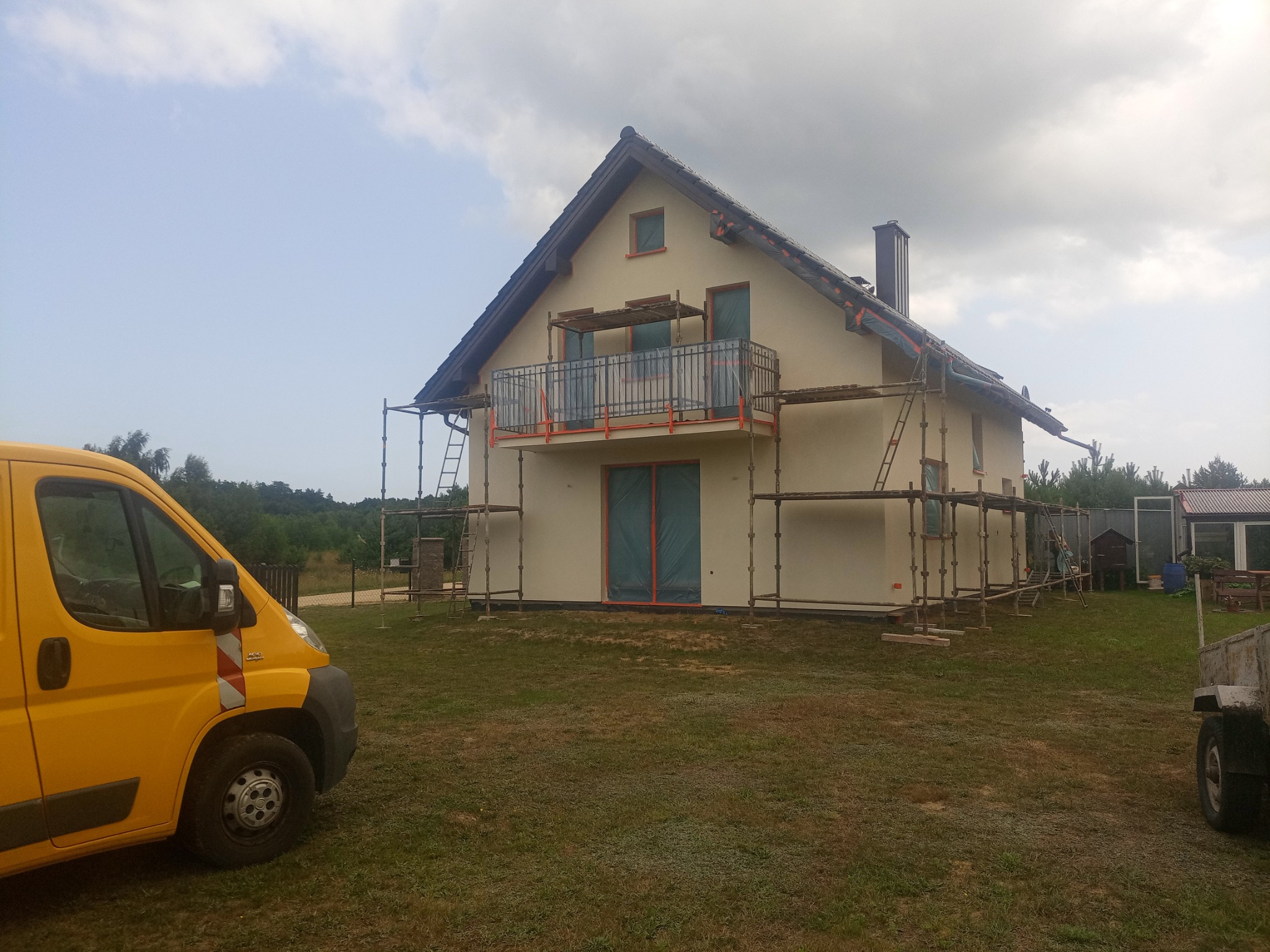 Dom jednorodzinny w trakcie prac elewacyjnych, z widocznym rusztowaniem, zabezpieczonymi oknami folią i żółtym samochodem dostawczym w tle.