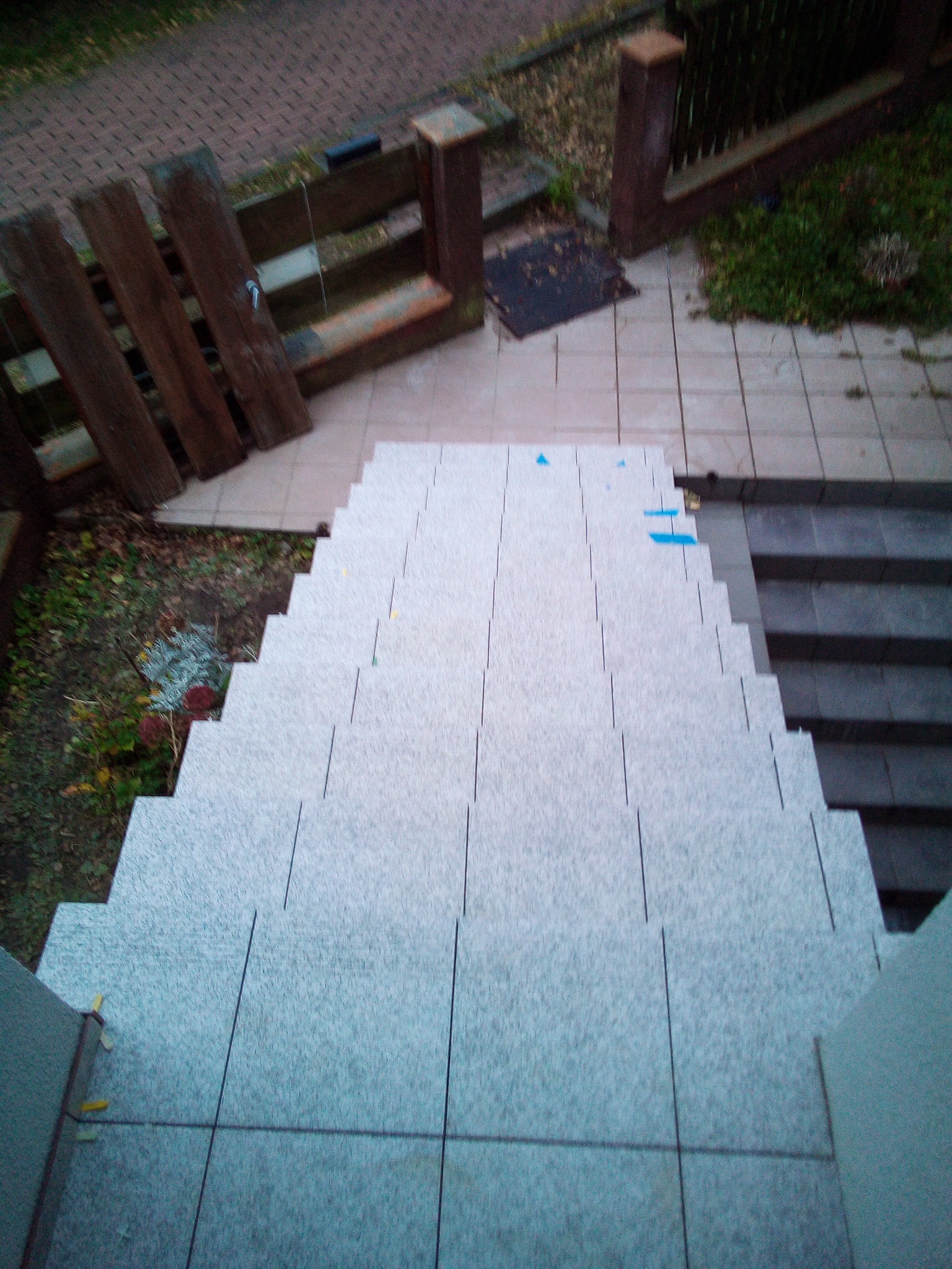 Wykończone schody zewnętrzne z szarych płytek, widok z góry z fragmentami niebieskiej taśmy zabezpieczającej na niektórych płytkach, obok widoczny fragment drewnianego ogrodzenia i roślinność.