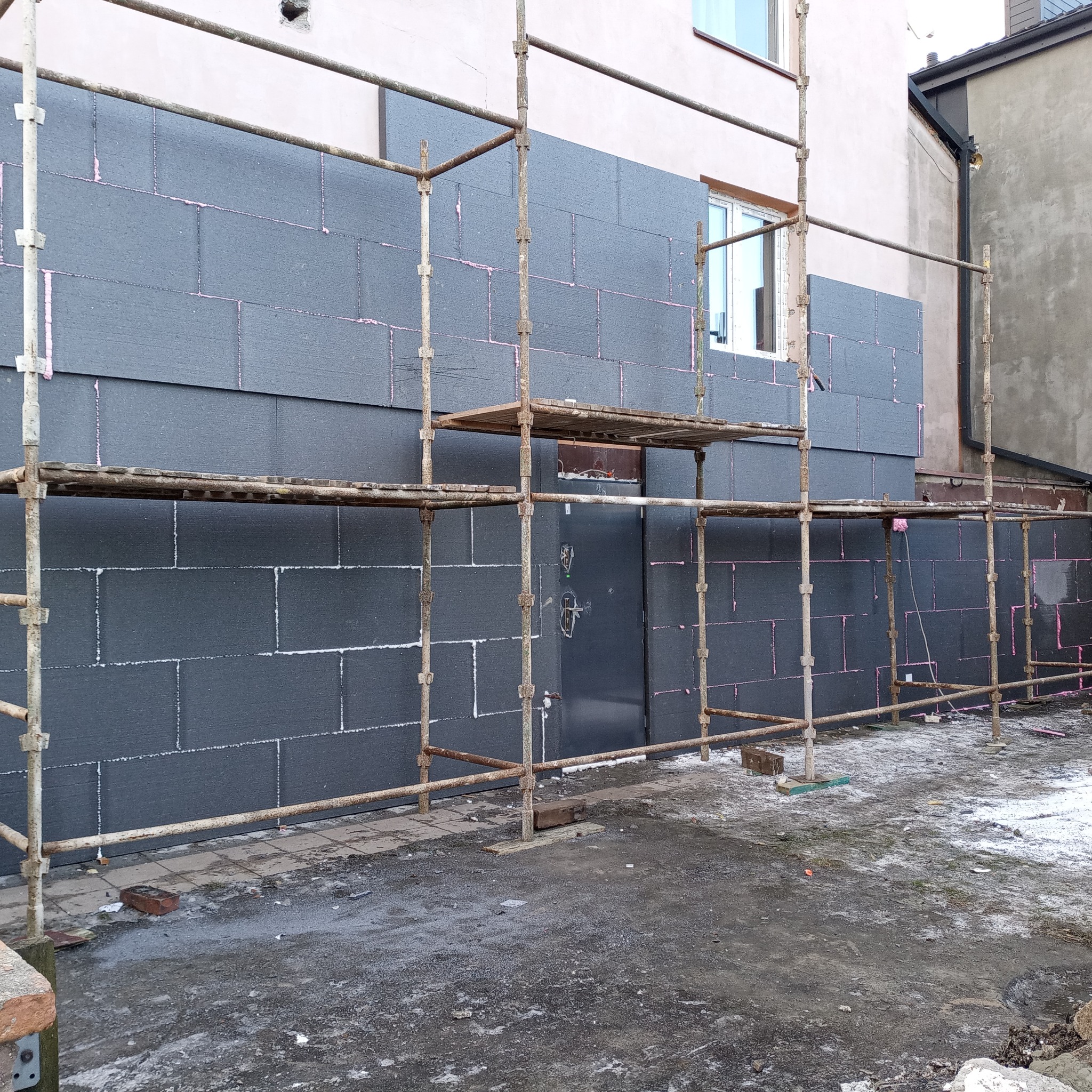 Montaż ciemnoszarej izolacji termicznej na elewacji budynku, widoczne rusztowanie i różowa pianka uszczelniająca między płytami.