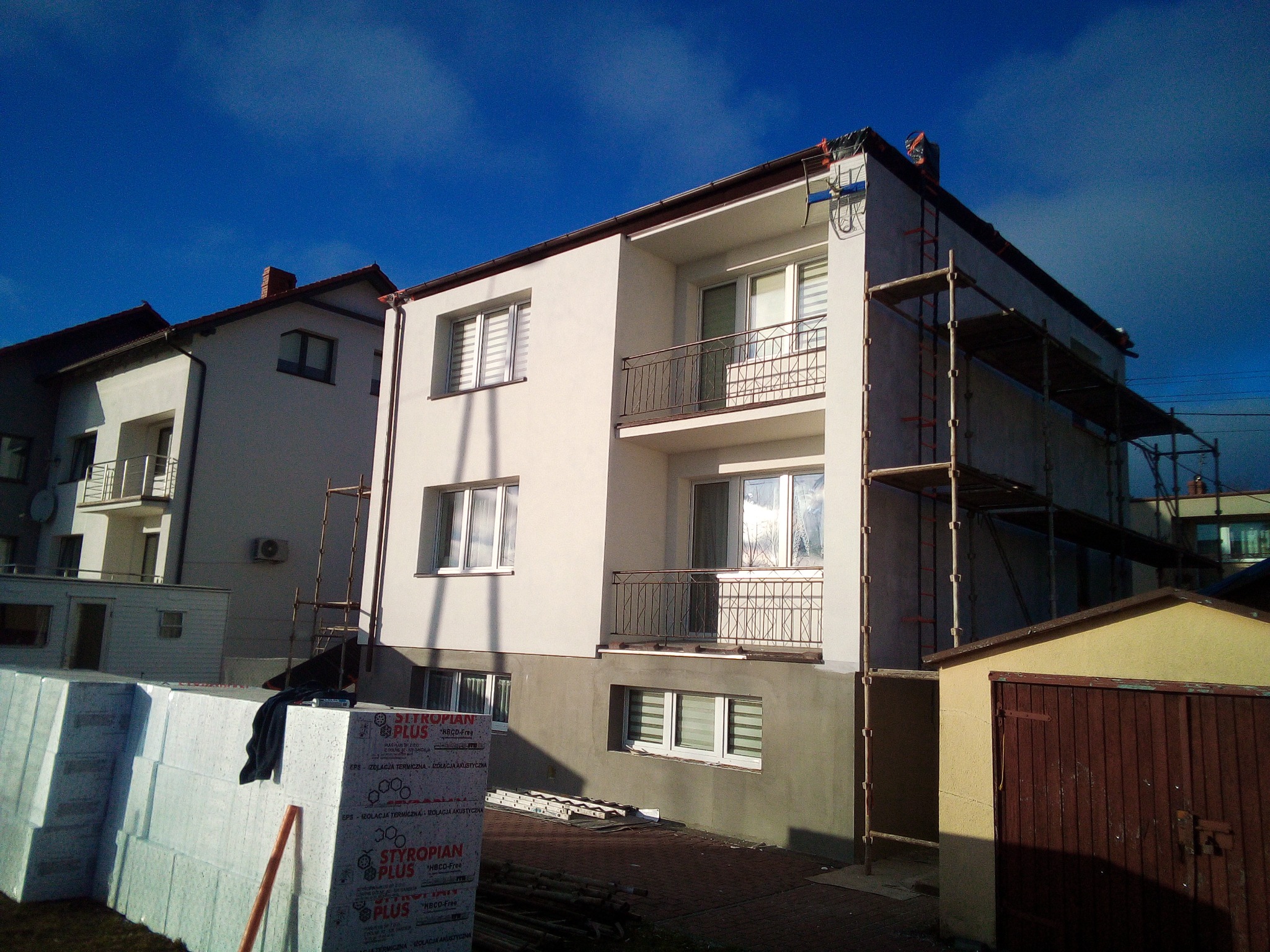Dwupiętrowy dom z balkonami w trakcie ocieplania elewacji, widoczne rusztowania, styropian i częściowo otynkowana dolna część budynku na tle błękitnego nieba.