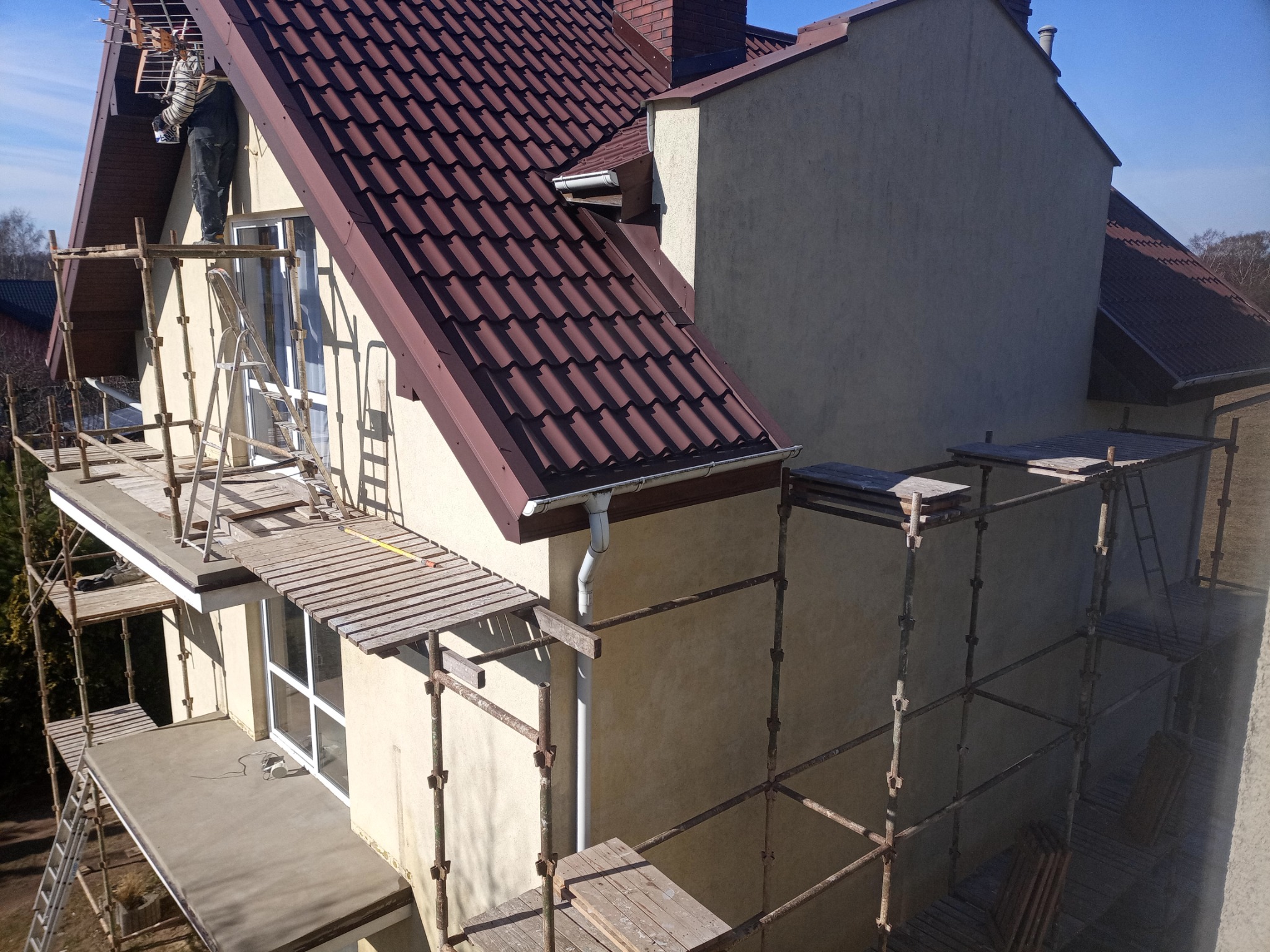 Praca na wysokości: mężczyzna na rusztowaniu przy elewacji domu z brązową dachówką, widoczny fragment okna i rynny, jasna elewacja w trakcie prac.