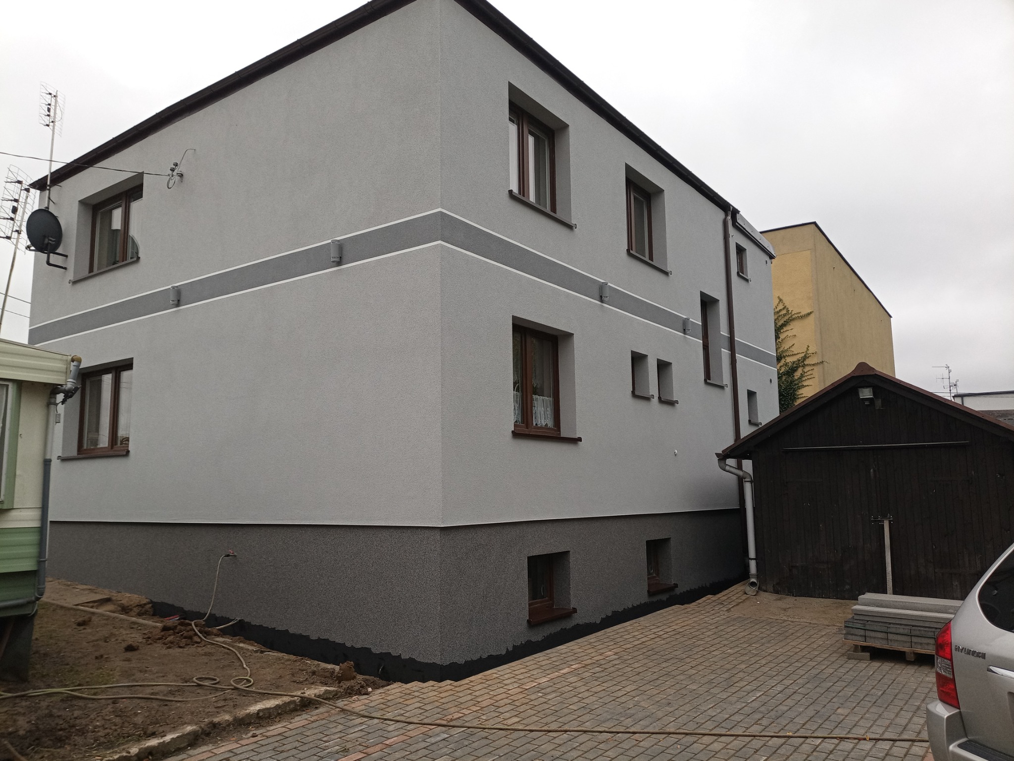 Dwukondygnacyjny dom z szarą elewacją i ciemniejszym cokołem, z widocznymi oknami z brązowymi ramami, ozdobiony poziomym pasem w jaśniejszym odcieniu szarości, z kostką brukową ułożoną przed...