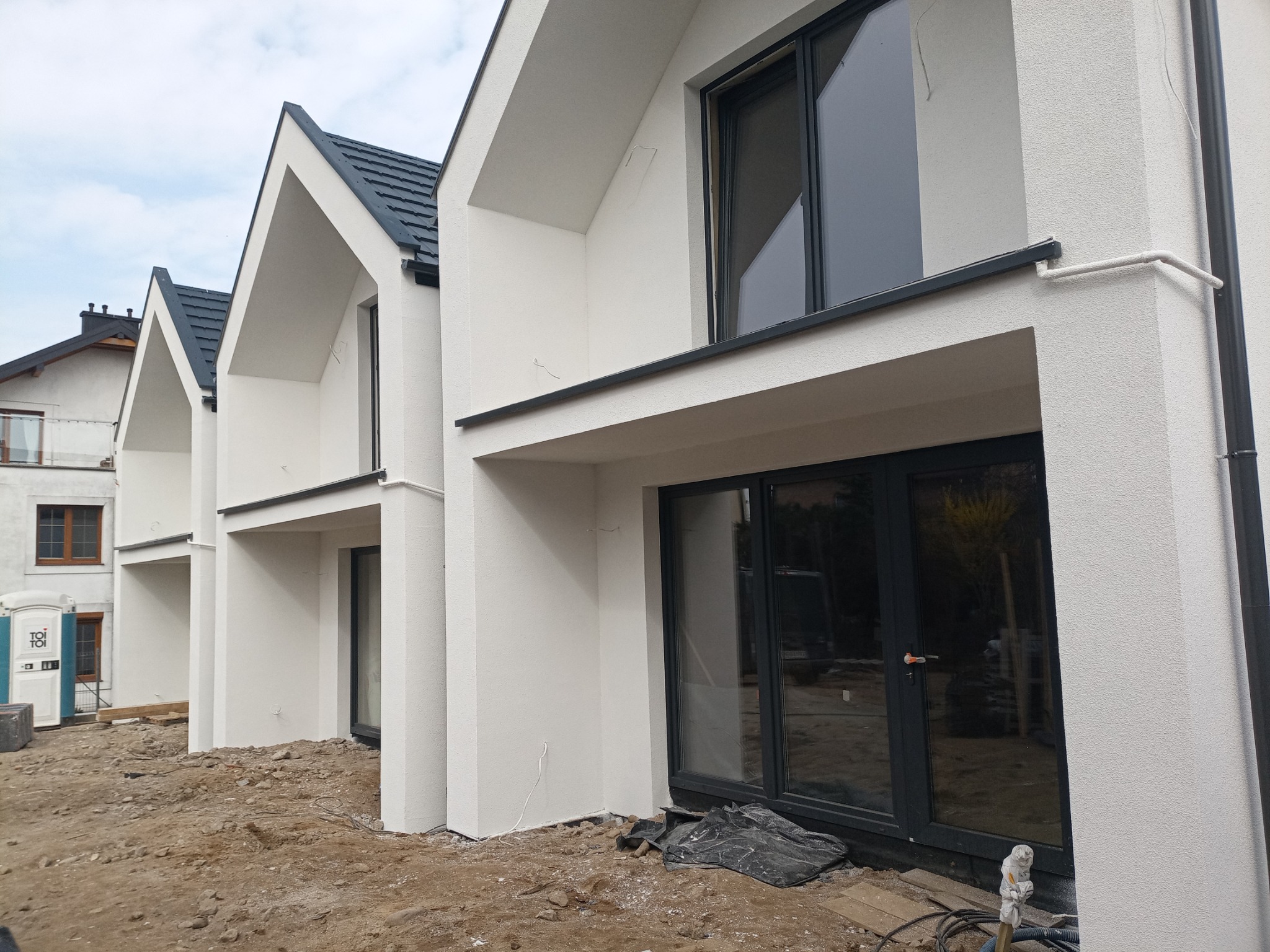 Nowoczesne domy w zabudowie szeregowej z białą elewacją o fakturze baranka, czarnymi oknami i grafitowym dachem, teren wokół w trakcie prac wykończeniowych.