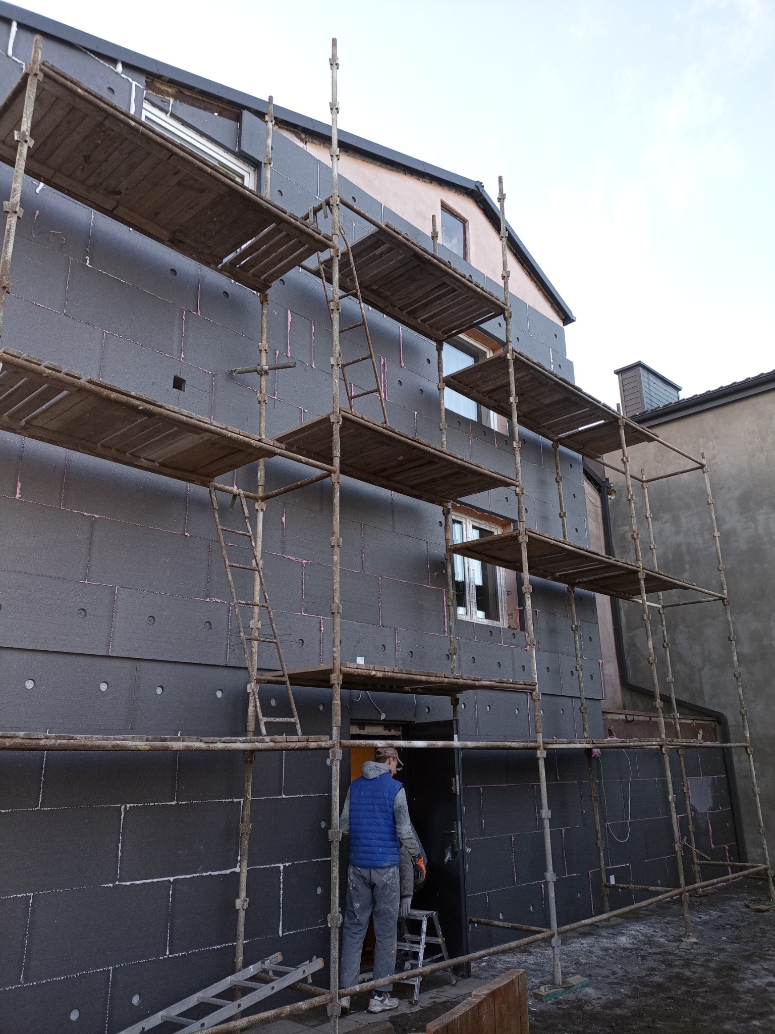 Budynek w trakcie ocieplania: szare płyty izolacyjne z różowymi kołkami mocowane do elewacji, rusztowanie z drewnianymi pomostami, pracownik w niebieskiej kamizelce na tle otwartych drzwi.