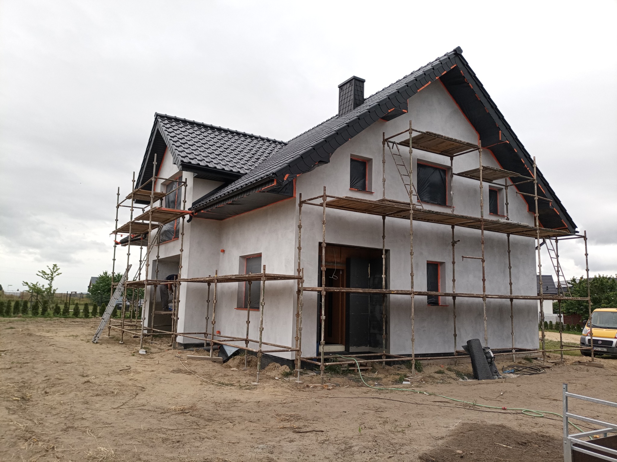 Dom jednorodzinny w trakcie ocieplania elewacji, widoczne rusztowania, czarna dachówka i okna zabezpieczone folią.