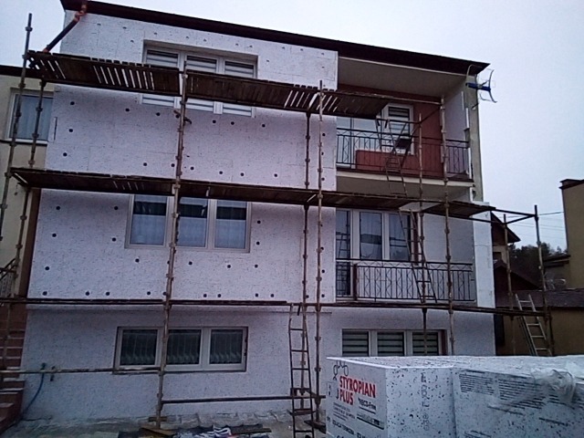 Dwupiętrowy dom w trakcie ocieplania elewacji, widoczne rusztowania, częściowo ocieplona ściana z widocznymi kołkami mocującymi styropian, na pierwszym planie paczki styropianu STYROPIAN J PLUS.