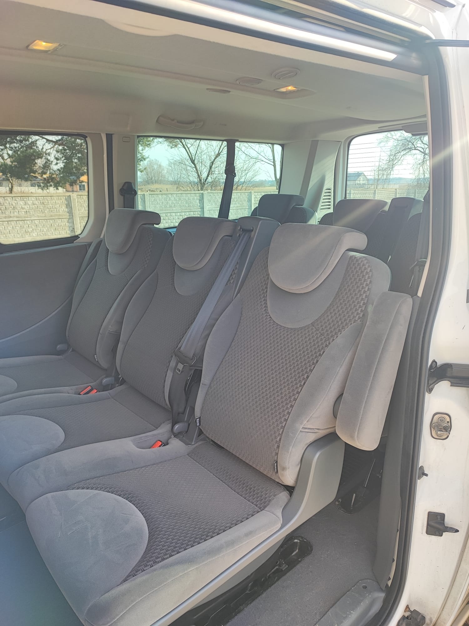 Wnętrze białego busa z trzema rzędami szarych foteli z pasami bezpieczeństwa, widok z otwartych drzwi na zewnątrz.