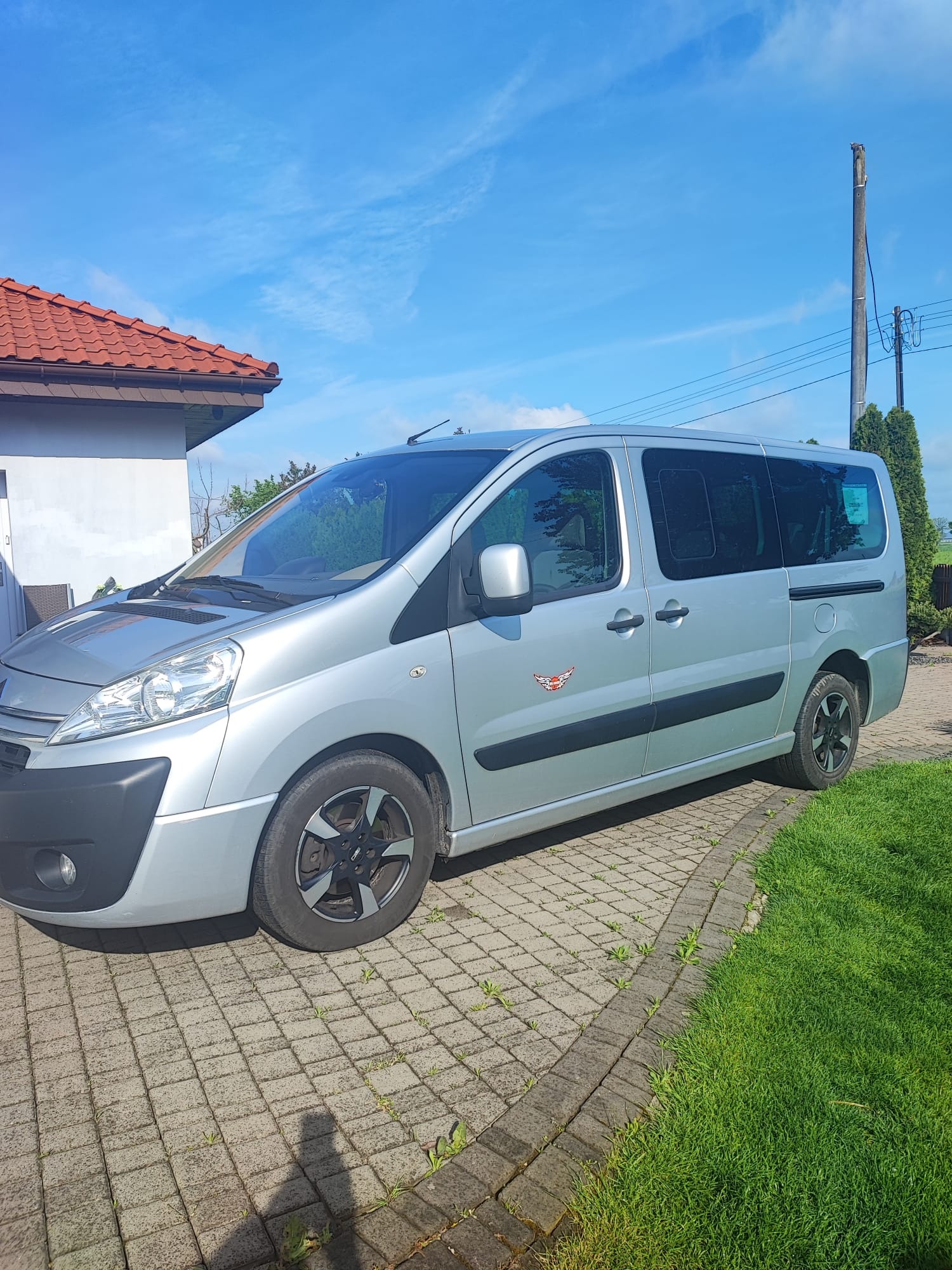 Srebrny van osobowy zaparkowany na kostce brukowej przed domem z czerwoną dachówką, widoczny z boku, z naklejką z motywem skrzydlatego orła na drzwiach.