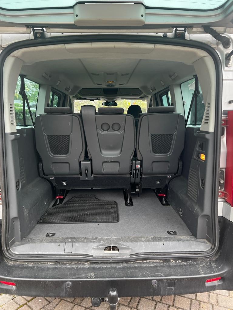 Wnętrze przestrzeni ładunkowej srebrnego vana z trzema rzędami foteli, widoczne z tyłu, z ciemną gumową wycieraczką na podłodze i hak holowniczy pod zderzakiem.