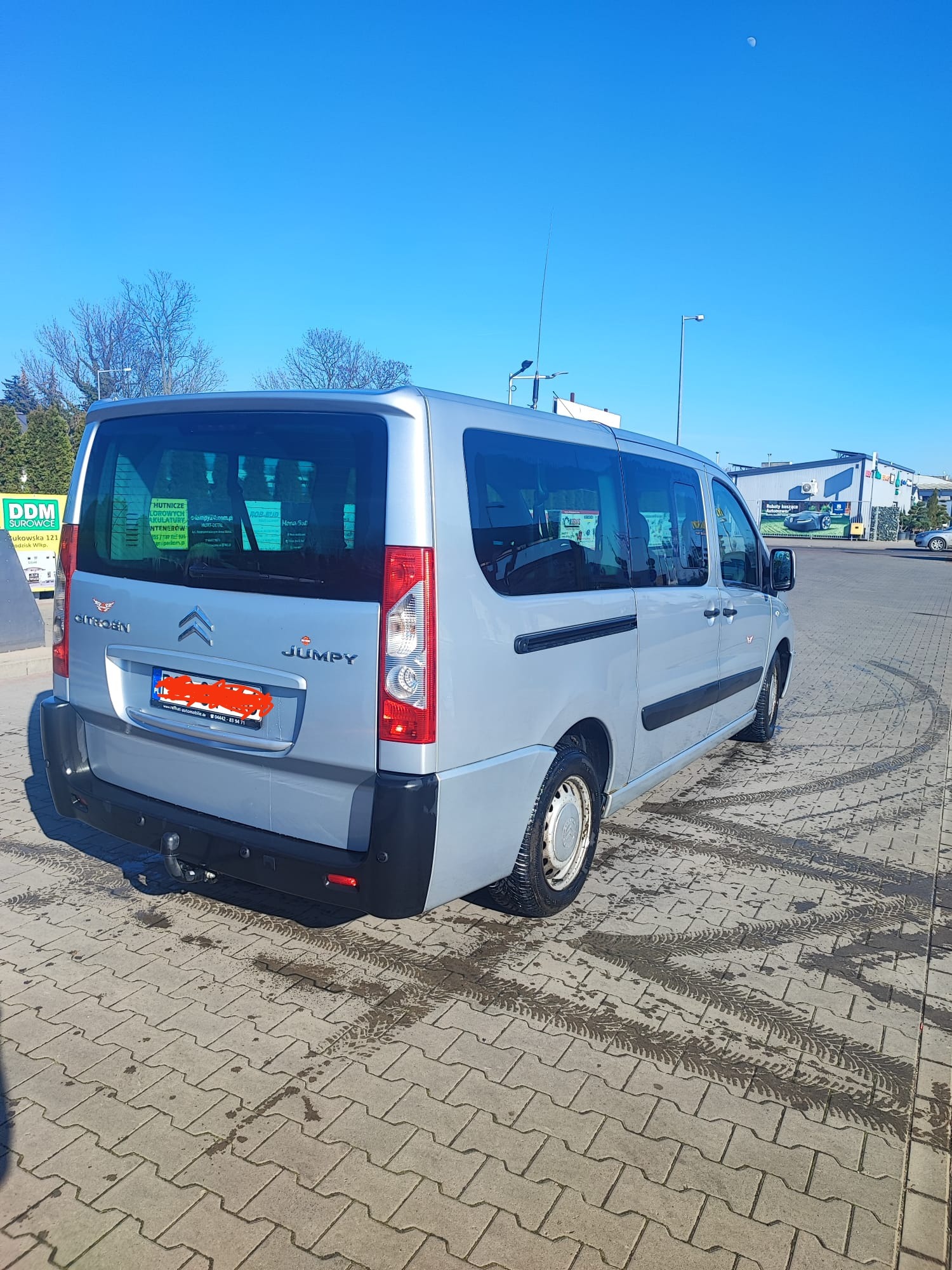 Srebrny Citroen Jumpy z hakiem holowniczym, zaparkowany na betonowej kostce brukowej z widocznymi śladami opon, błękitne niebo w tle.