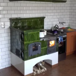 Zielony piec kaflowy z paleniskiem, z zapasem drewna pod spodem, w kuchni wyłożonej białymi płytkami.