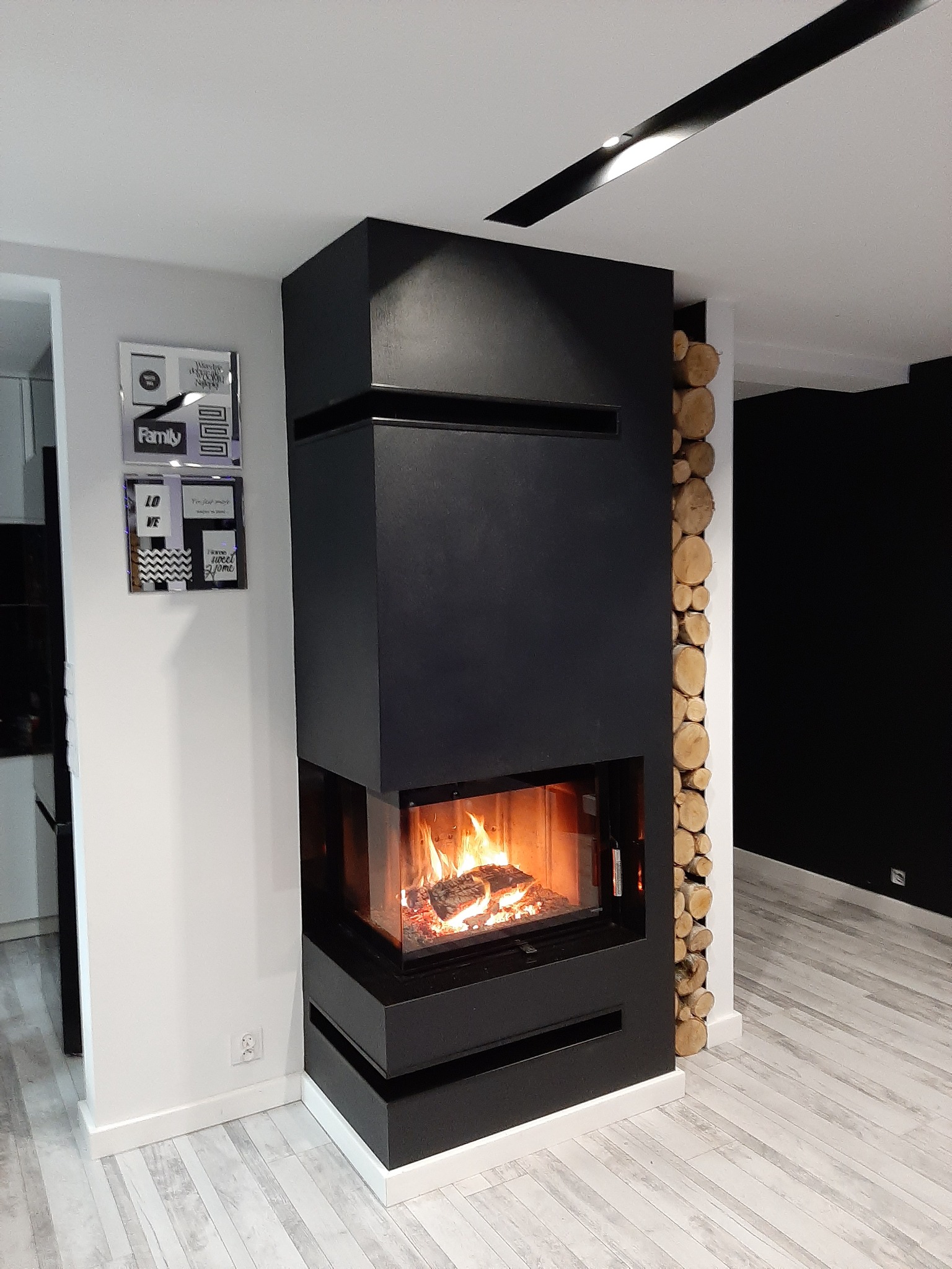 Nowoczesny, czarny piec wolnostojący z paleniskiem, otoczony dekoracyjnym obłożeniem z drewna, w minimalistycznym wnętrzu z drewnianą podłogą i czarną ścianą w tle.
