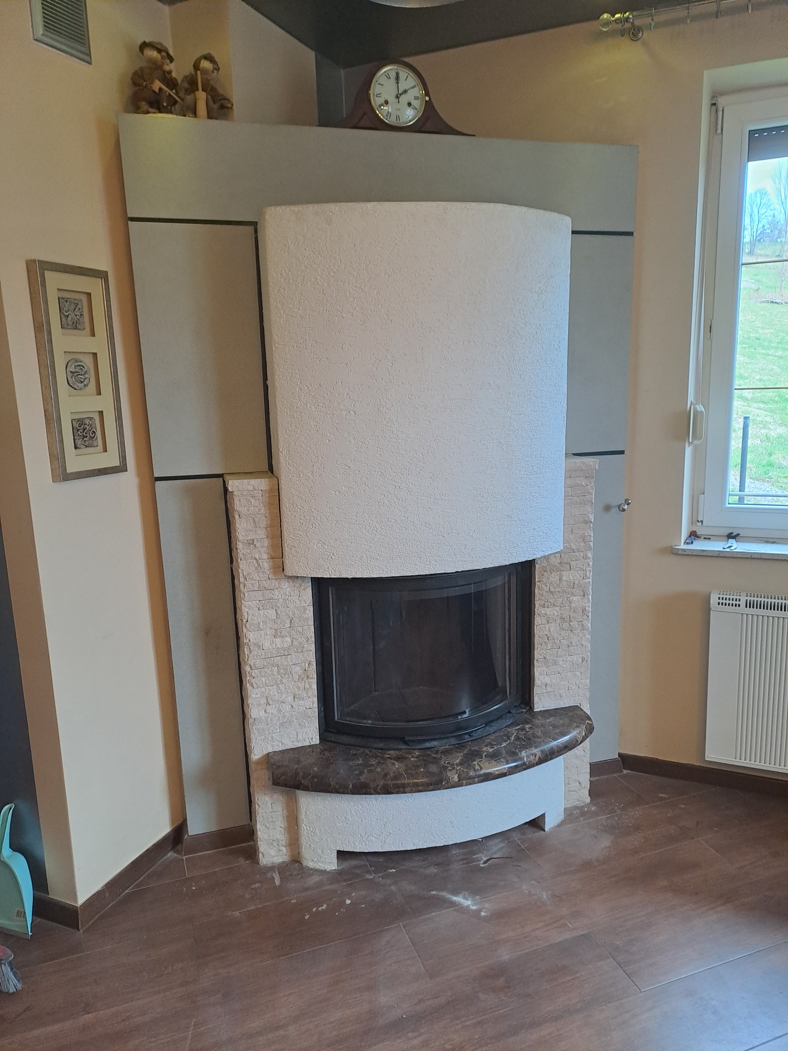 Obudowa wkładu kominkowego z białego, chropowatego tynku, obłożona kamieniem dekoracyjnym, z blatem z ciemnego marmuru, w salonie z drewnopodobną podłogą.