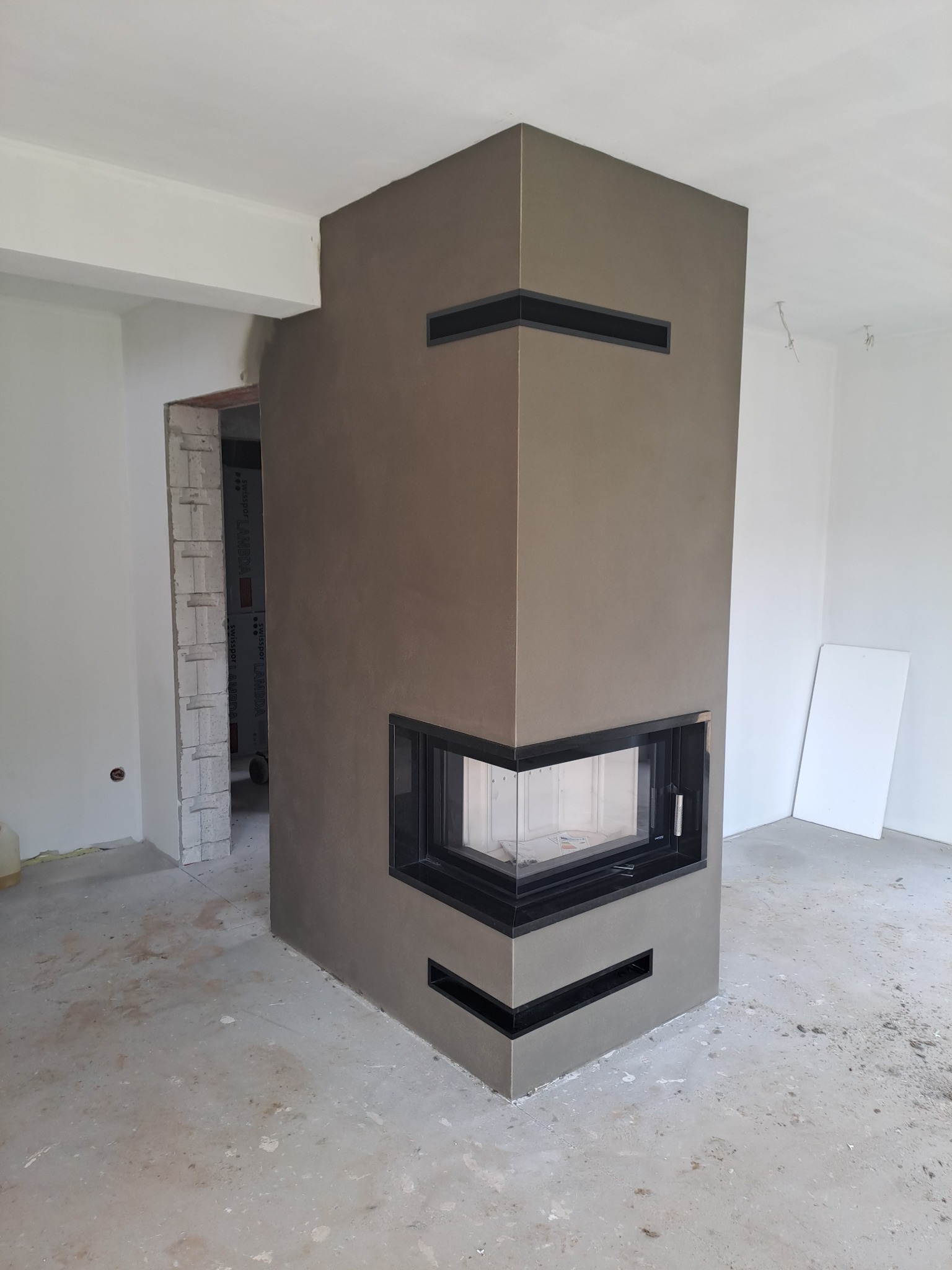 Nowoczesny, narożny kominek obudowany szarym tynkiem strukturalnym z czarnymi, poziomymi wstawkami wentylacyjnymi, w surowym wnętrzu w trakcie wykańczania.