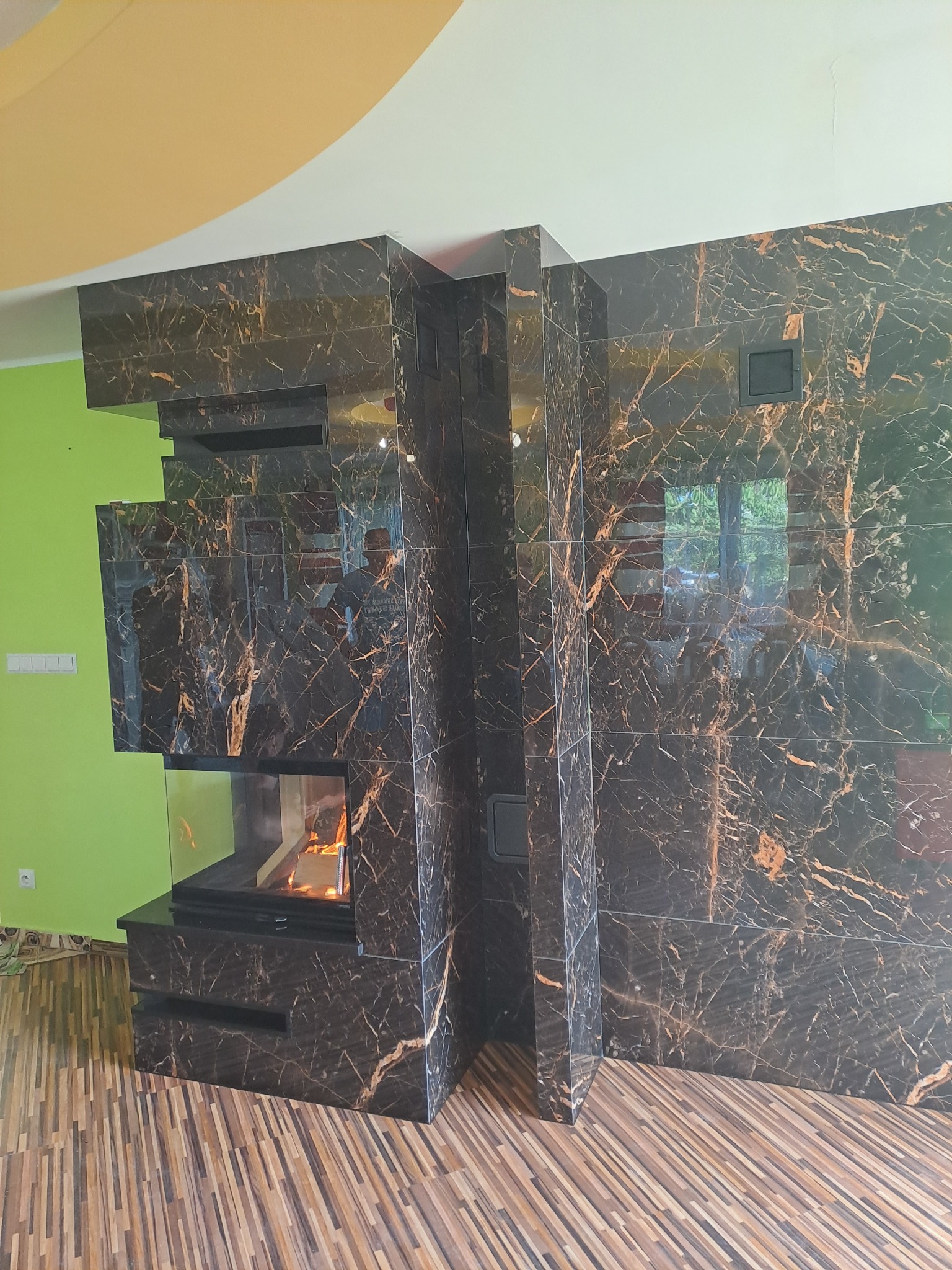 Nowoczesny wkład kominkowy z czarnym marmurem z wyraźnym, złotym użyleniem, zabudowany w ścianie z płyt marmurowych, z widocznym paleniskiem i płomieniami.