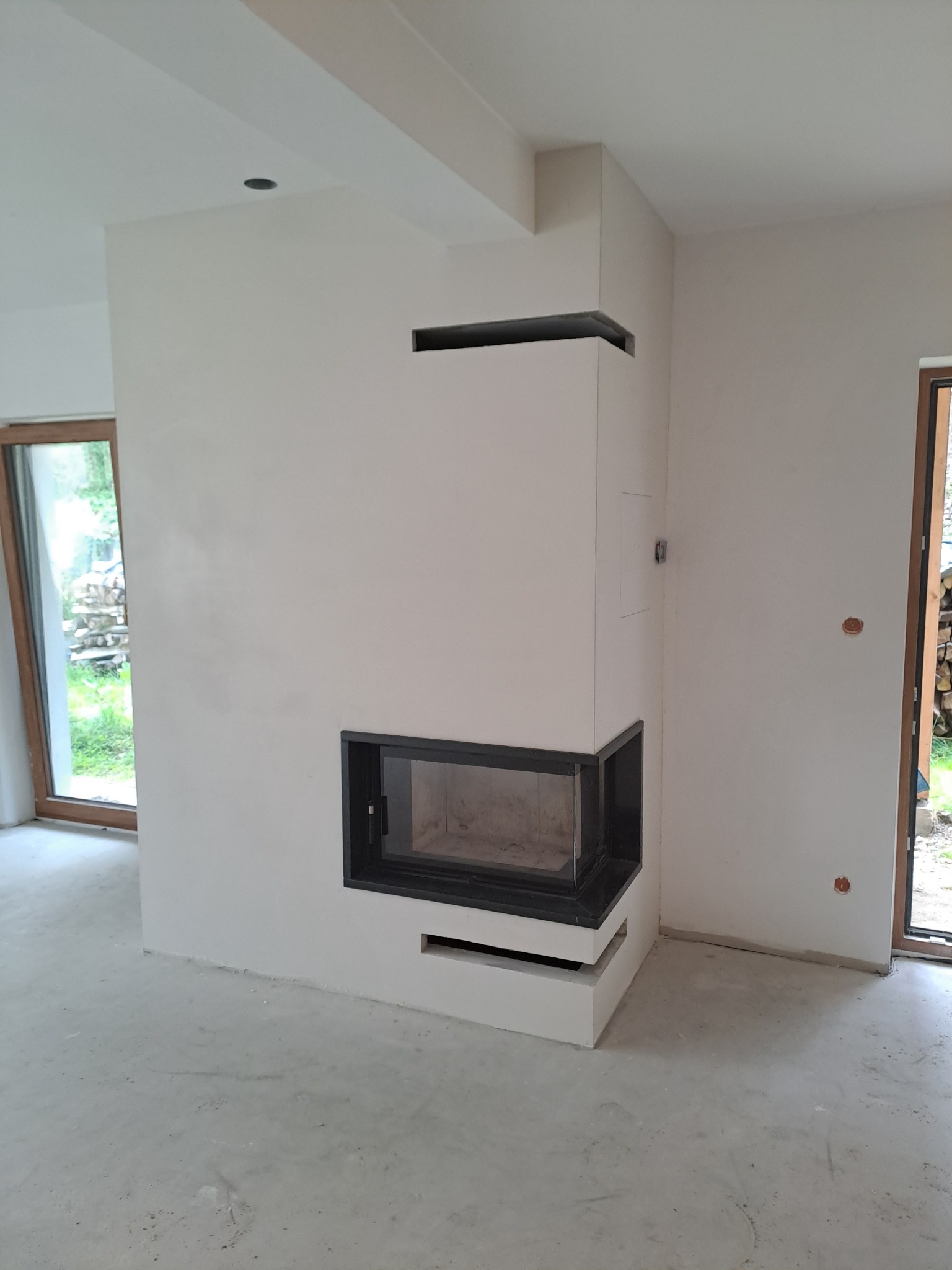 Nowoczesny narożny wkład kominkowy z czarną ramą, obudowany jasnym tynkiem, widoczny w surowym wnętrzu z oknami i betonową podłogą.