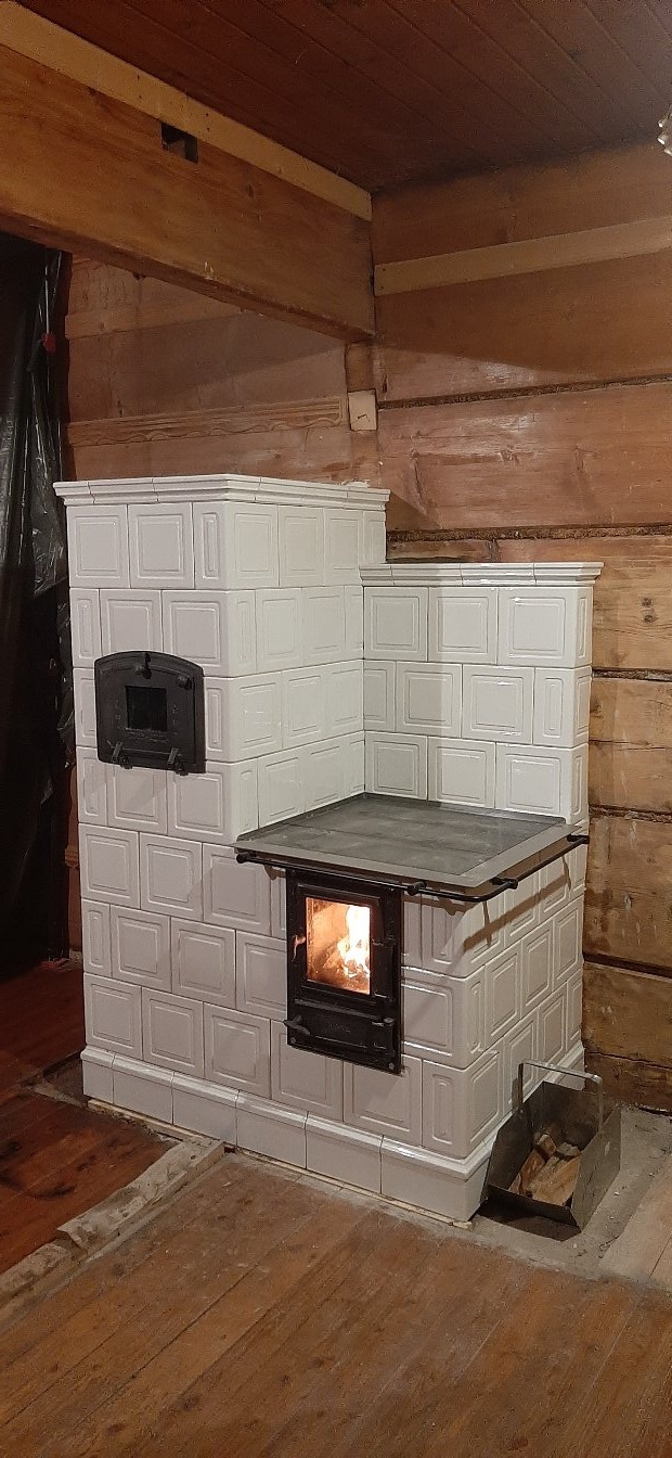 Biały piec kaflowy w rustykalnym wnętrzu z drewnianymi ścianami, z paleniskiem, w którym widać płomienie.