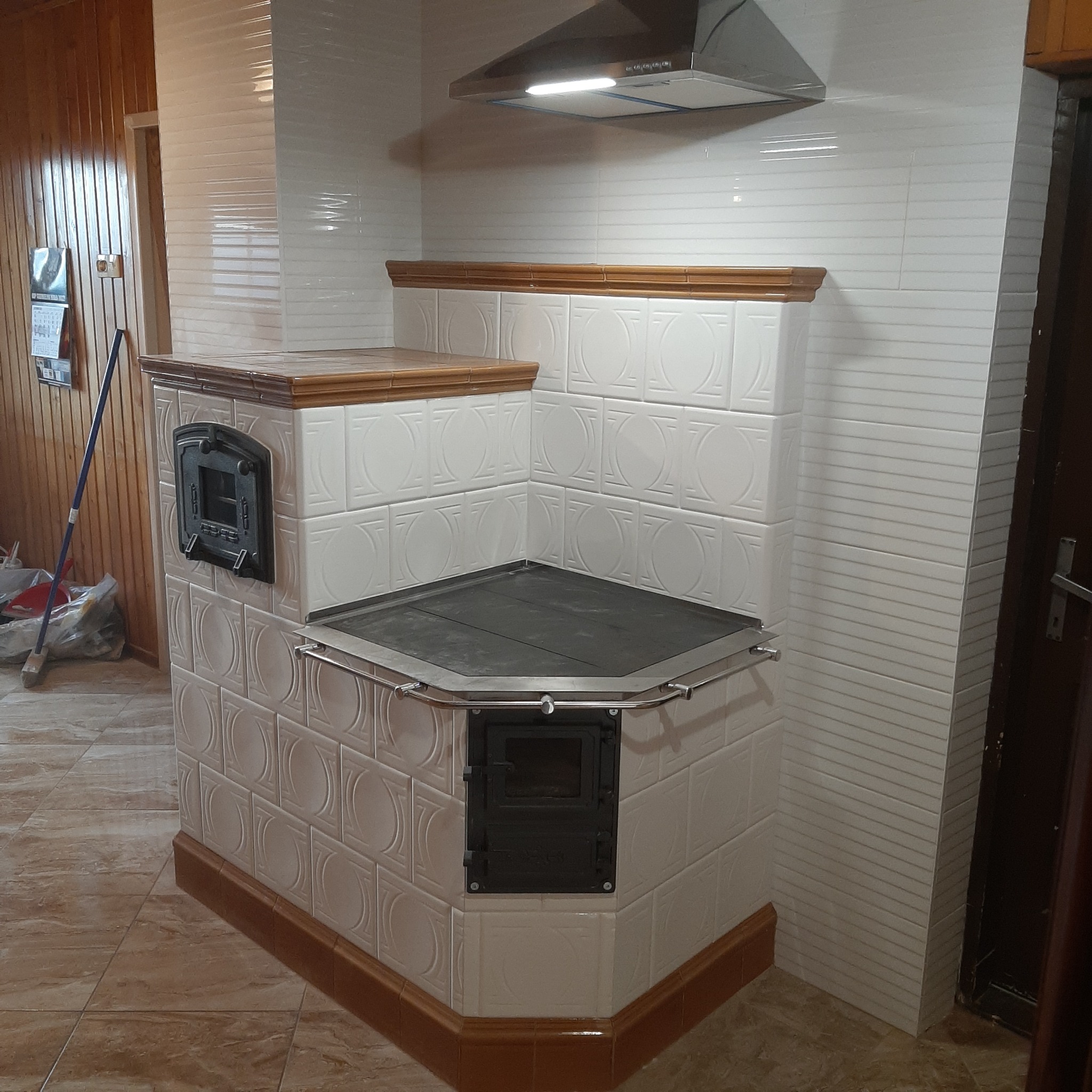 Biały piec kaflowy z dwoma czarnymi drzwiczkami i metalową poręczą, zwieńczony drewnianym blatem, pod okapem w kuchni z białymi płytkami.