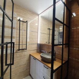 KABINETMEBLE TOMASZ LENCZEWSKI - Nowoczesna łazienka z umywalką z kamienia na drewnianym blacie i białą szafką, oddzielona czarną, metalową ramą. Beżowe płytki na ścianach i podświetlane lustro.