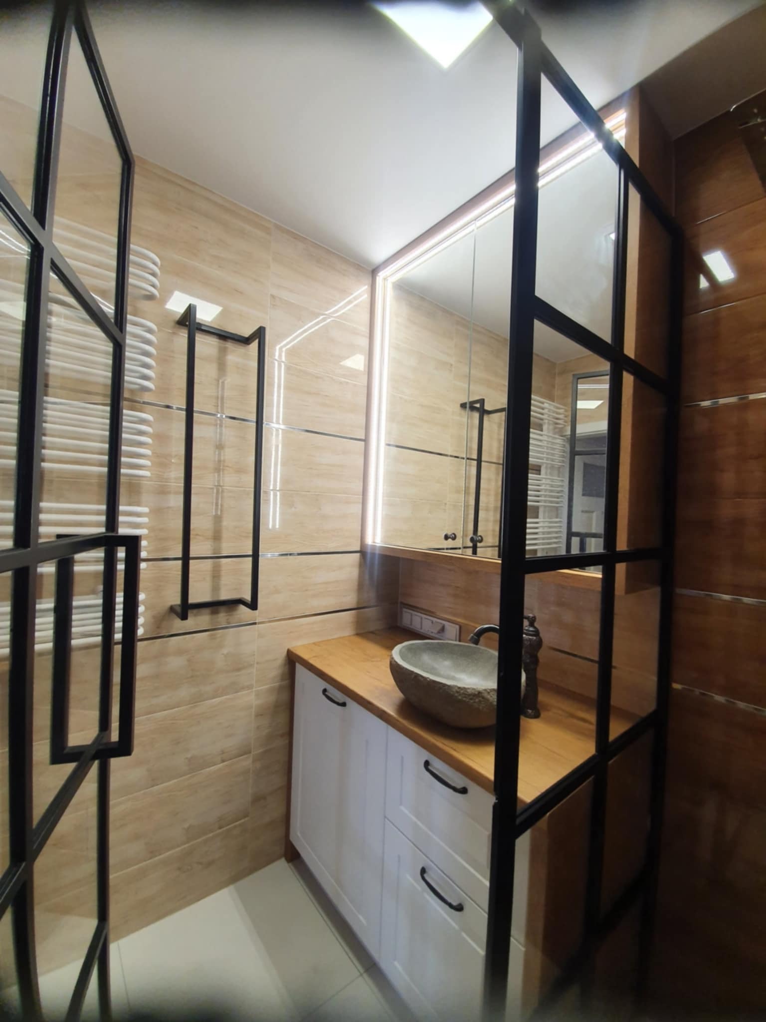 Nowoczesna łazienka z umywalką z kamienia na drewnianym blacie i białą szafką, oddzielona czarną, metalową ramą. Beżowe płytki na ścianach i podświetlane lustro.