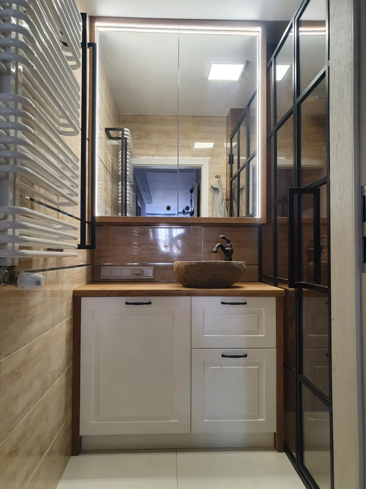 Nowoczesna łazienka z szafką pod umywalkę z kamienia, lustrem z podświetleniem LED i ścianką działową w stylu loft. Umywalka w formie kamiennej misy.