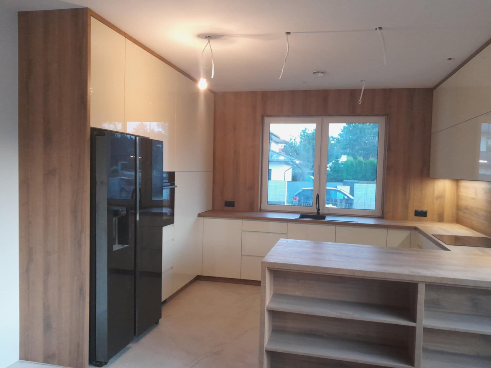 Nowoczesna kuchnia z białymi, lakierowanymi frontami i drewnianymi akcentami. Widoczna wyspa kuchenna i duża lodówka. Oświetlenie w trakcie montażu.